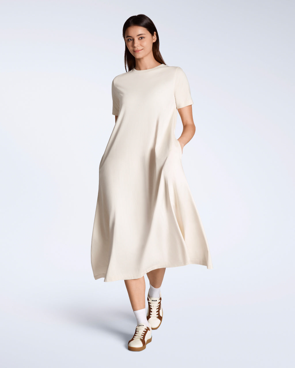 A woman smiling into the camera, walking, with her hands in her pockets modelling a plain natural coloured, long line, maxi organic cotton t shirt dress.