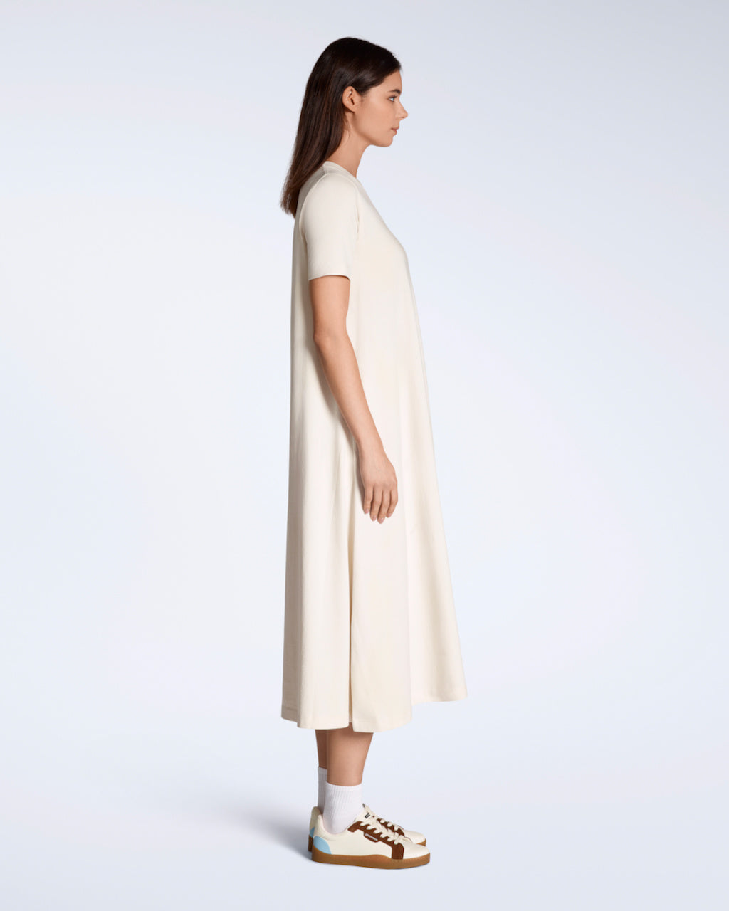 A woman facing side on with her arms by her side, modelling a plain natural coloured, long line, maxi organic cotton t shirt dress.