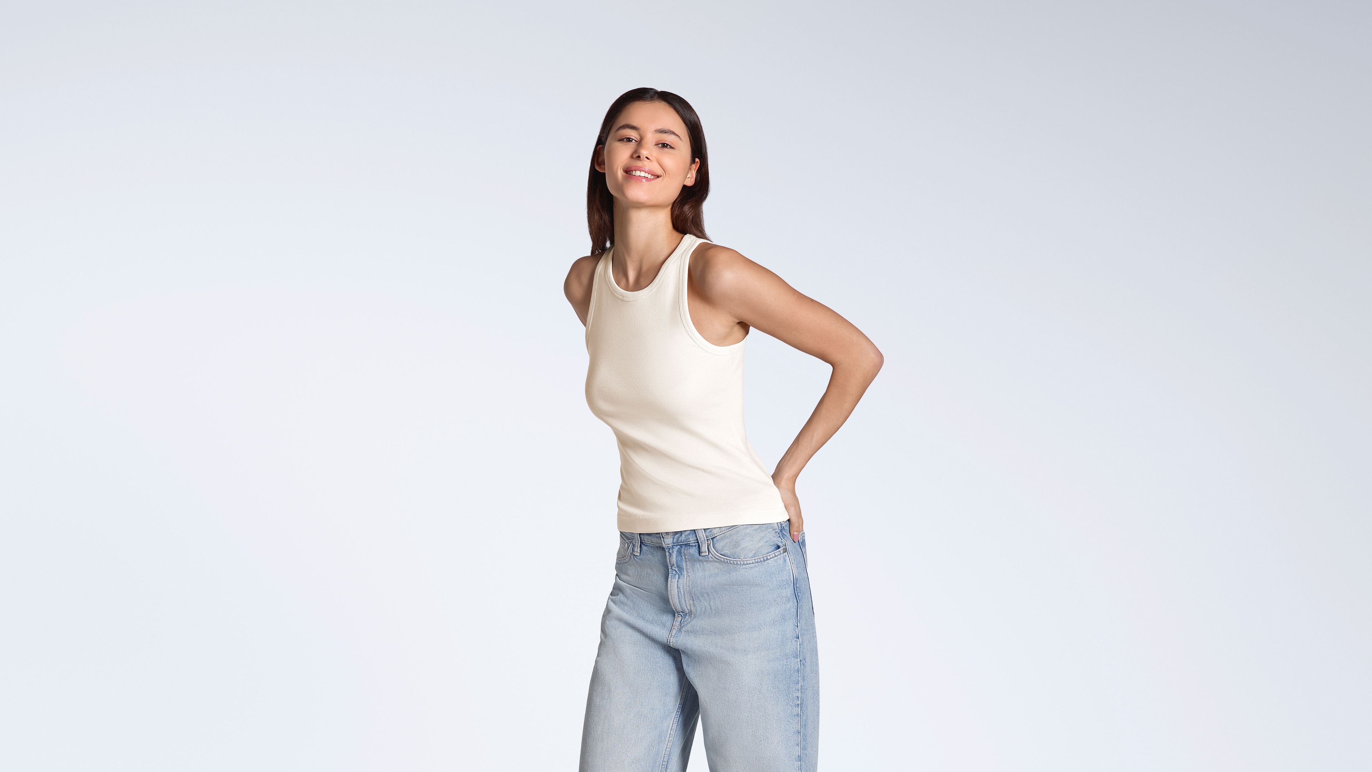 A woman smiling into the camera, modelling a plain natural coloured, organic cotton tank top with a pair of jeans.