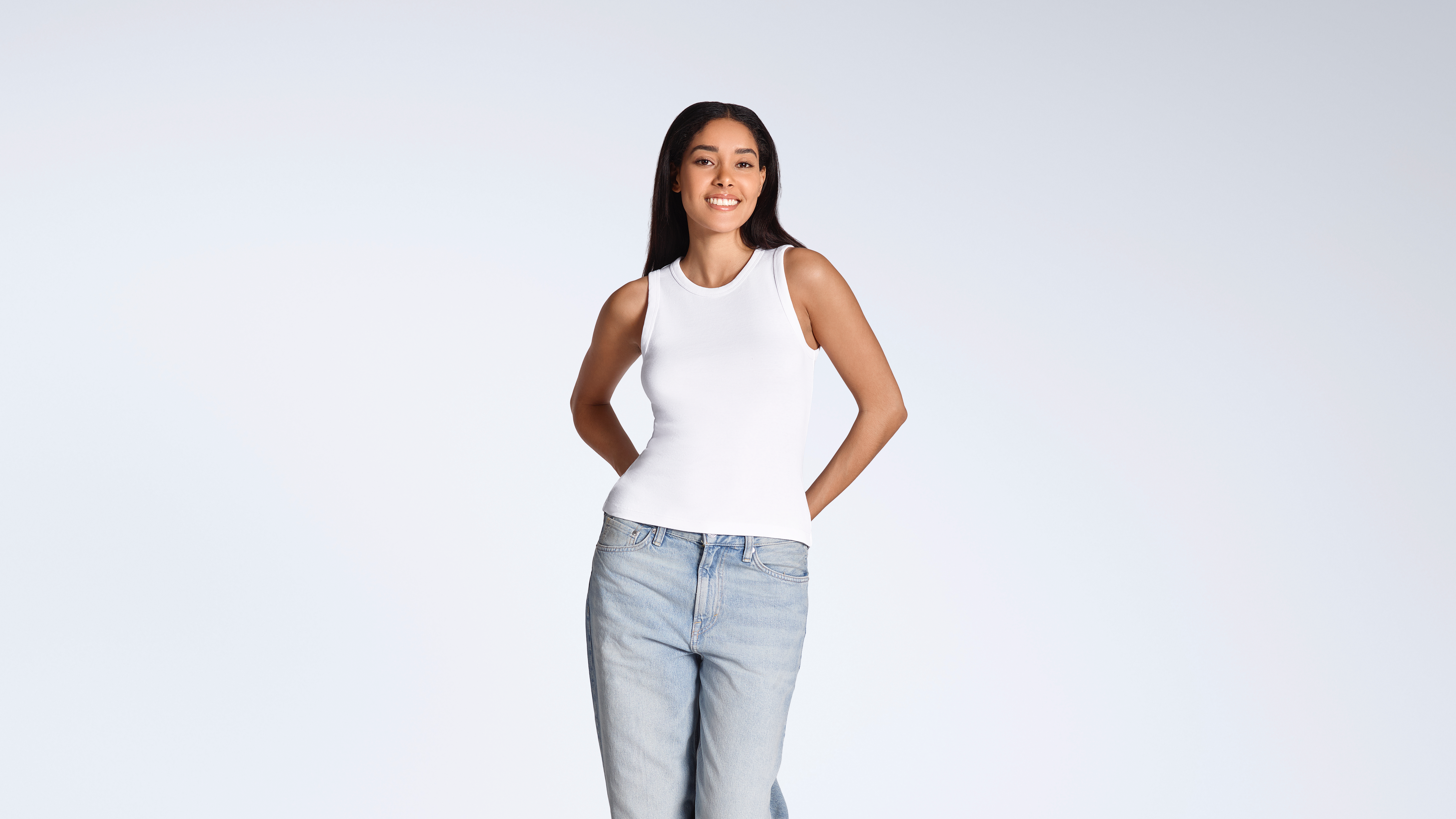 A woman smiling into the camera, modelling a plain white, organic cotton tank top with a pair of jeans.