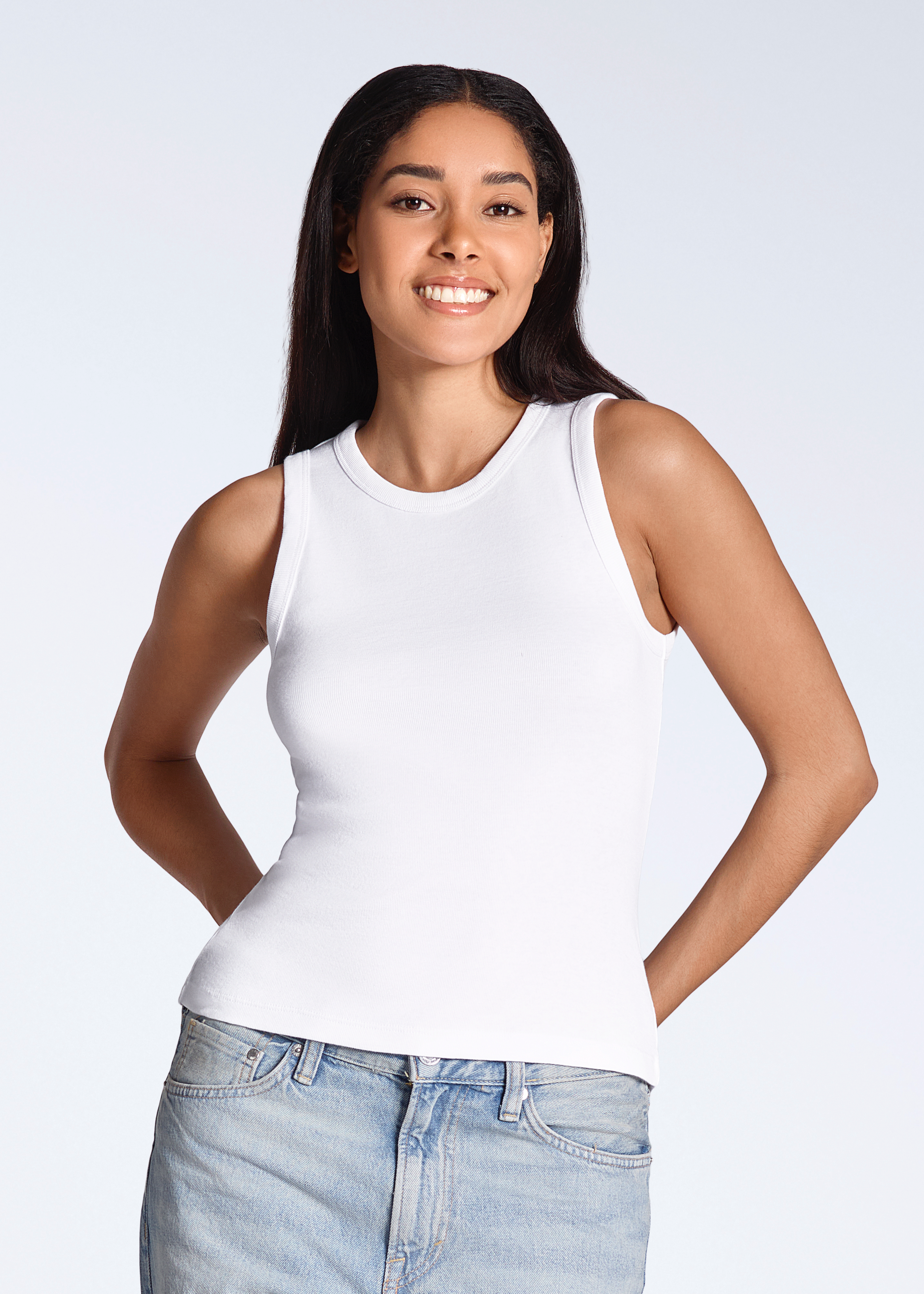 A woman smiling into the camera, modelling a plain white, organic cotton tank top with a pair of jeans.