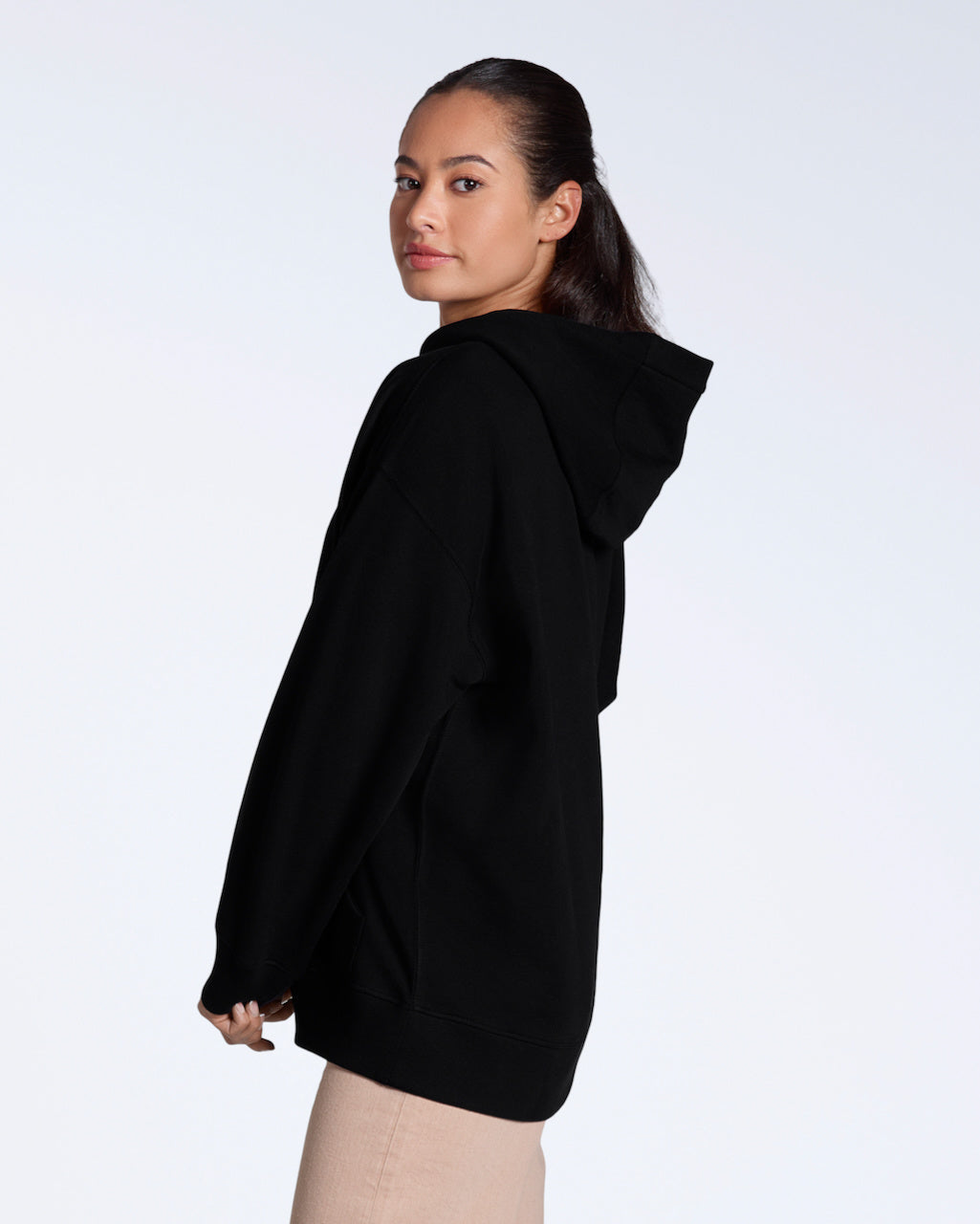 A woman facing side on looking back at the camera, modelling a plain black, oversized, organic cotton hoodie.