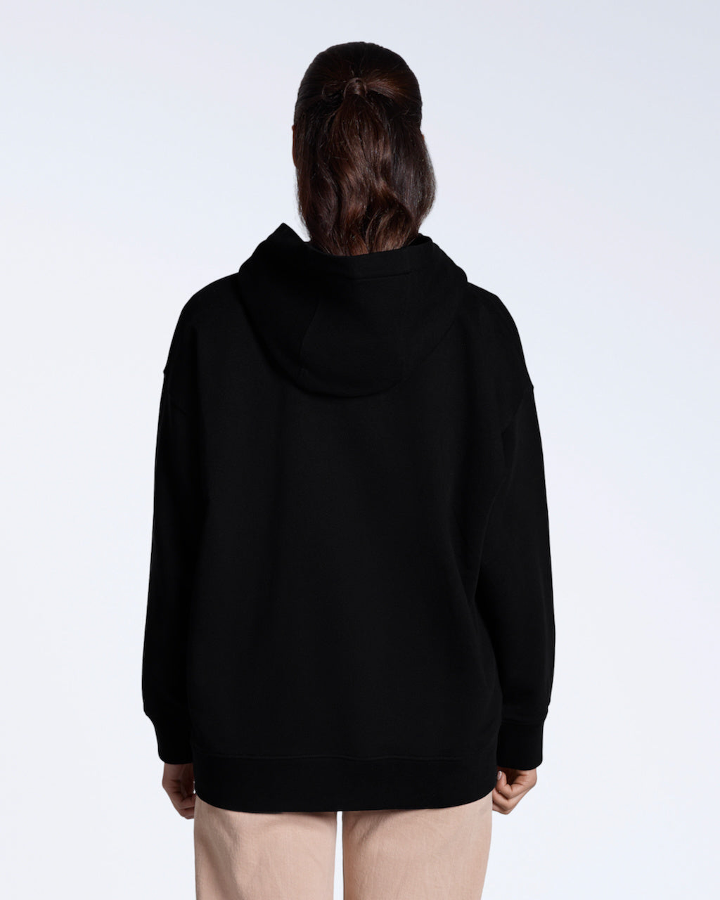 A woman facing away from the camera with her hands by her sides, modelling a plain black, oversized, organic cotton hoodie.