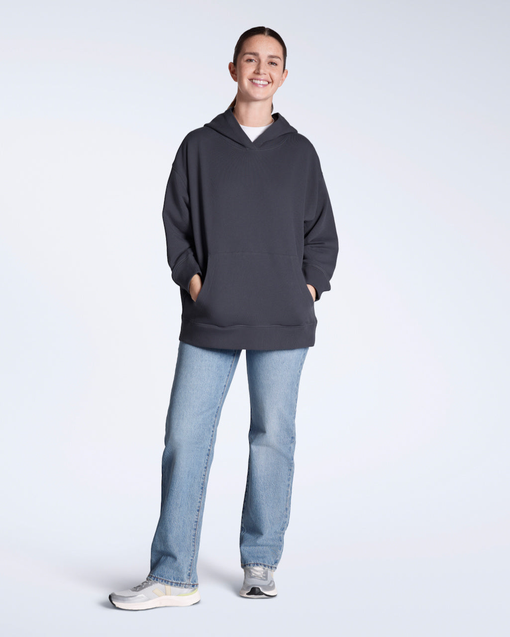 A full body photo of a woman smiling into the camera with her hands in her pockets, modelling a plain dark grey, oversized, organic cotton hoodie with a pair of blue jeans.