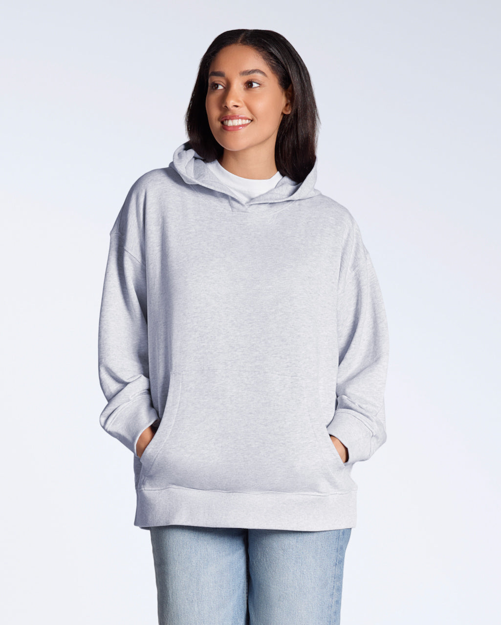 A woman smiling looking away from the camera with her hands in her pockets, modelling a plain grey, oversized, organic cotton hoodie.