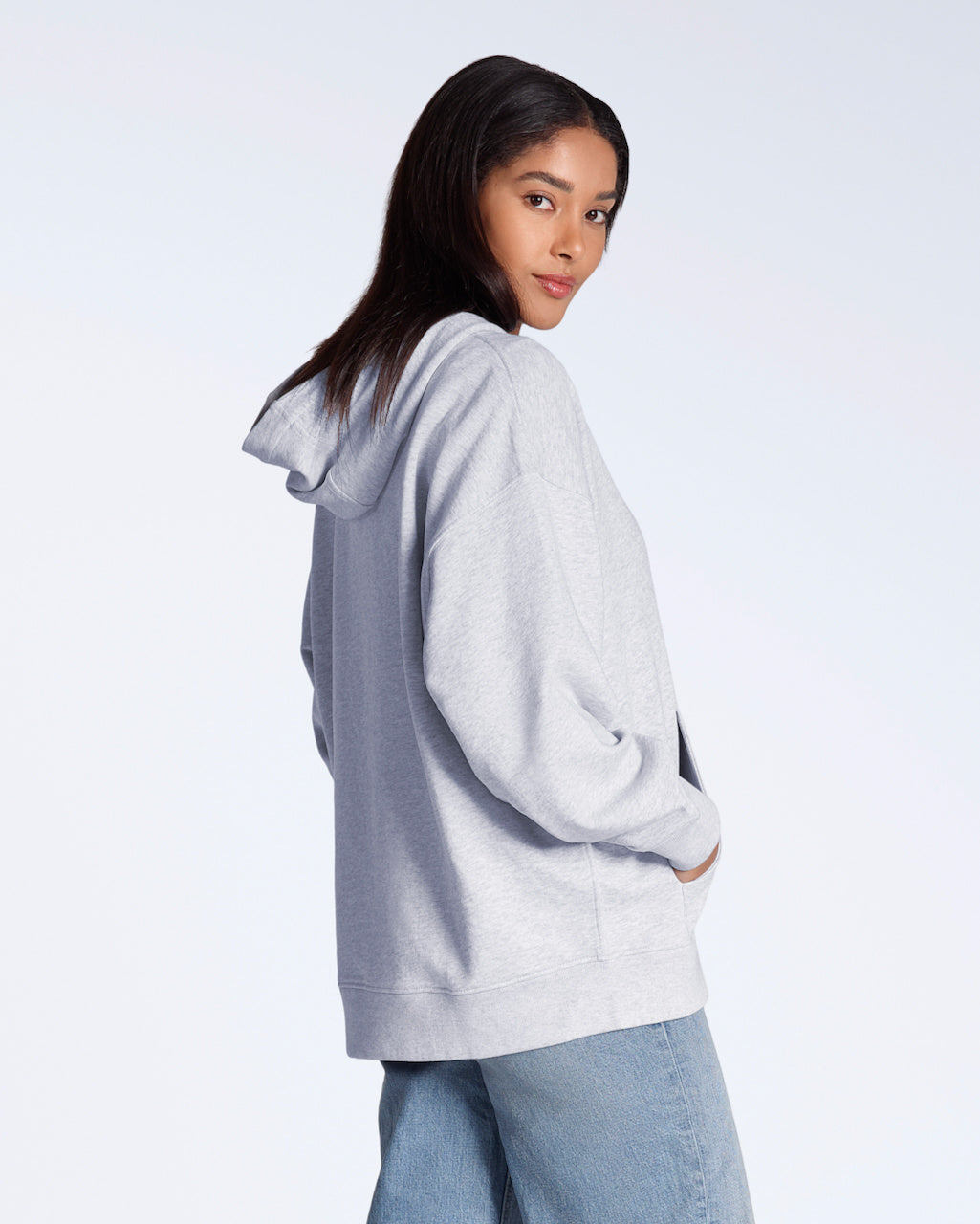 A woman facing side on looking back at the camera, modelling a plain grey, oversized, organic cotton hoodie.