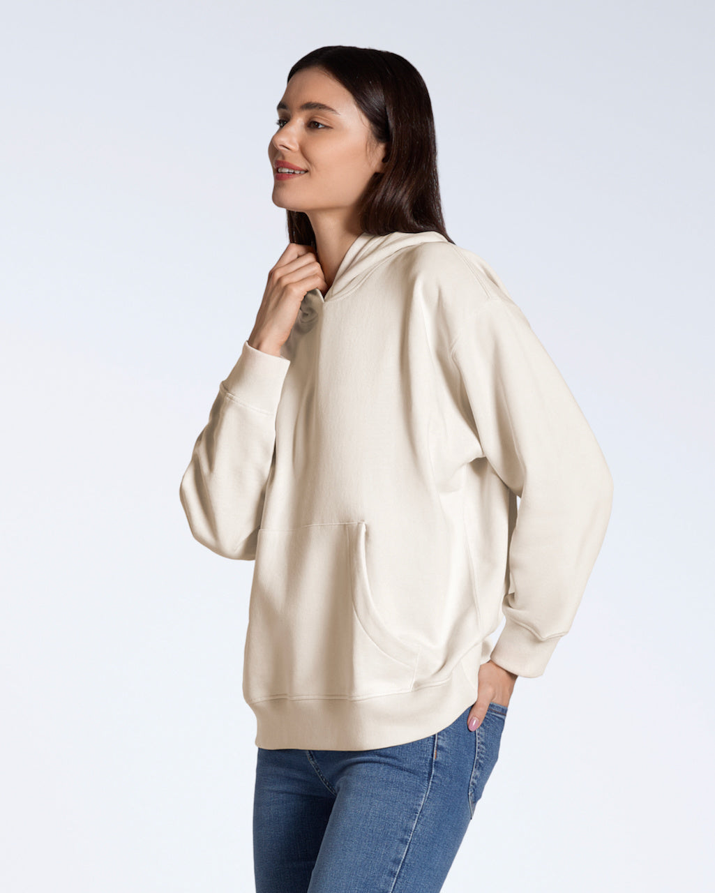 A woman smiling with her hand in her back pocket, modelling a natural coloured, oversized, organic cotton hoodie.