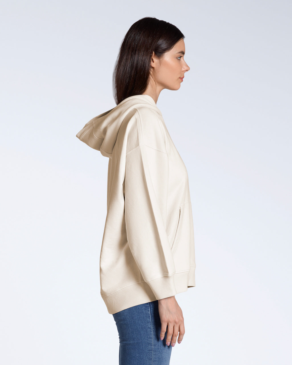 A woman facing side on, modelling a plain natural coloured, oversized, organic cotton hoodie.