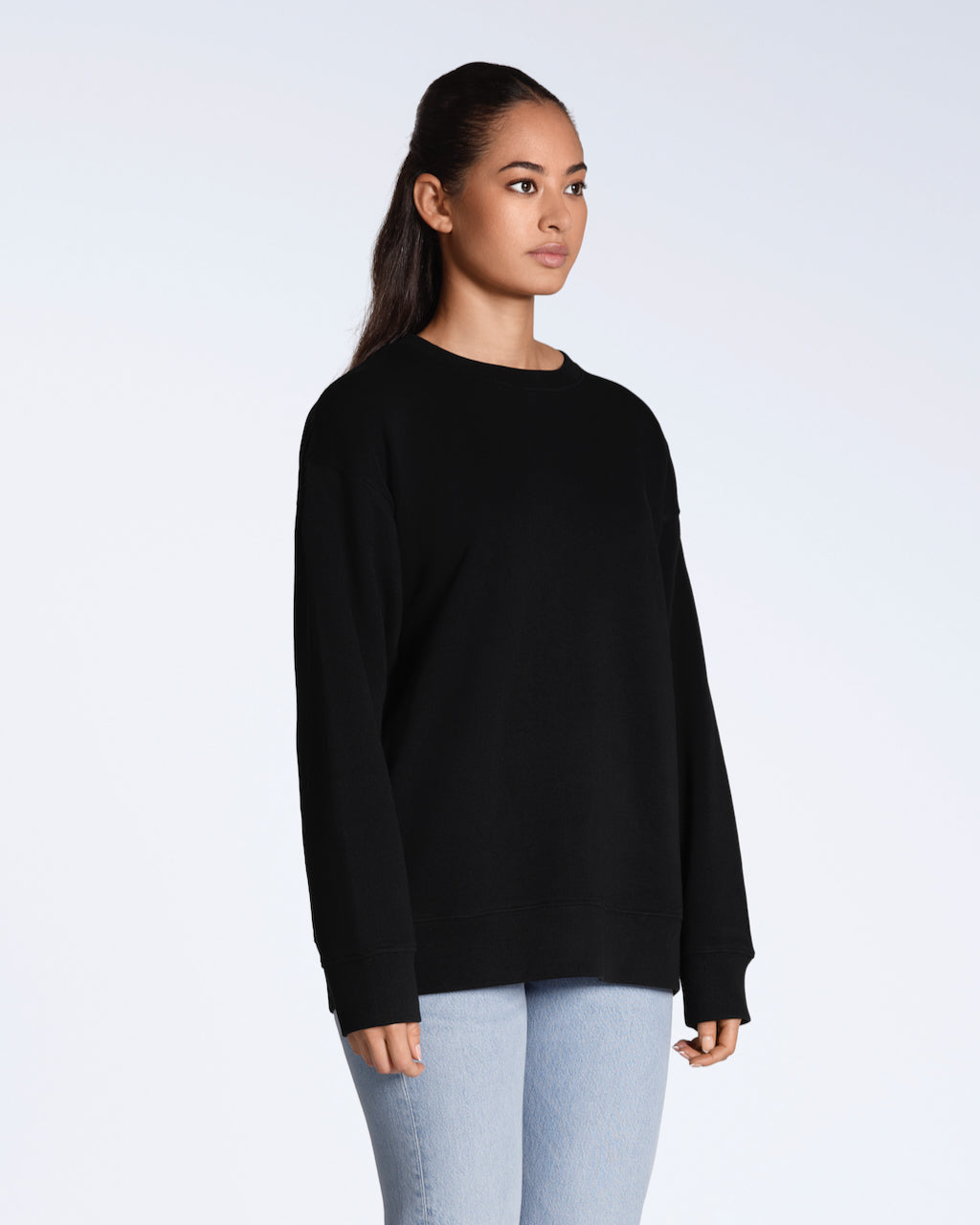 A woman looking away from the camera at an angle with her arms by her side, modelling a plain black, oversized, organic cotton sweatshirt.