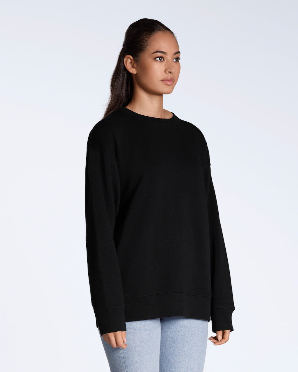 A woman looking away from the camera at an angle with her arms by her side, modelling a plain black, oversized, organic cotton sweatshirt.