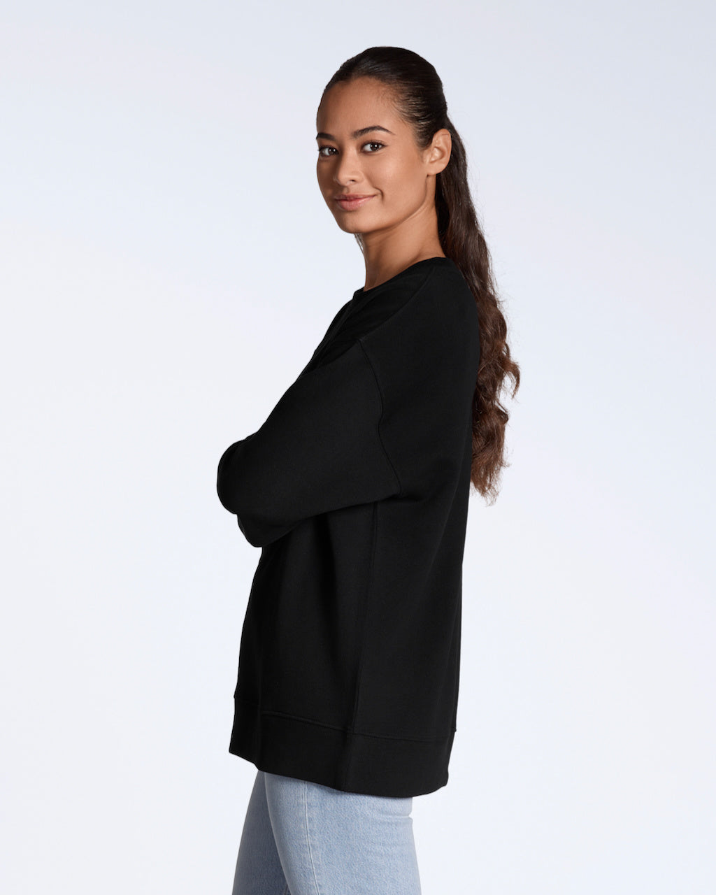 A woman facing side on smiling back at the camera, with her arms crossed, modelling a plain black, oversized, organic cotton sweatshirt.