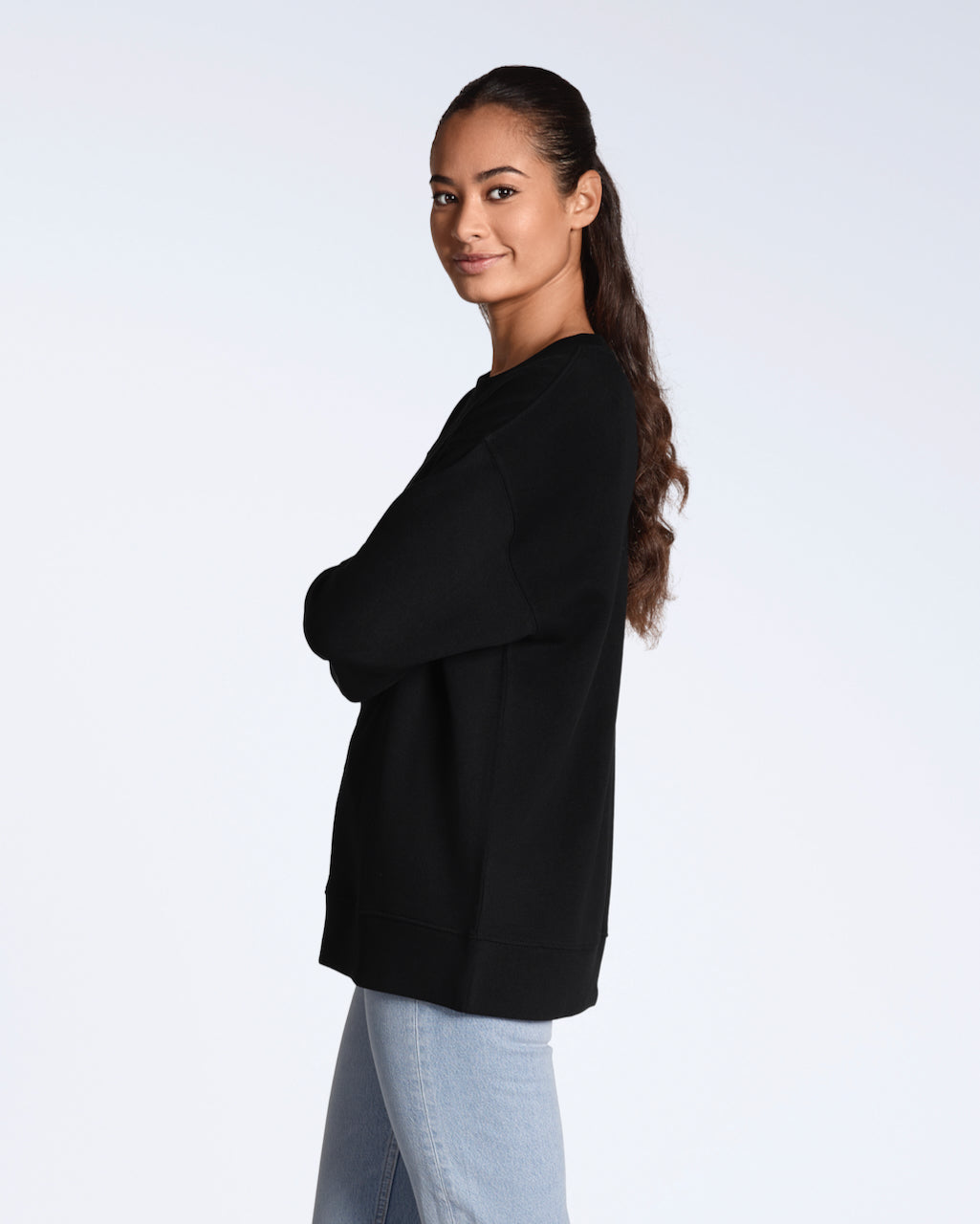 A woman facing side on smiling back at the camera, with her arms crossed, modelling a plain black, oversized, organic cotton sweatshirt.