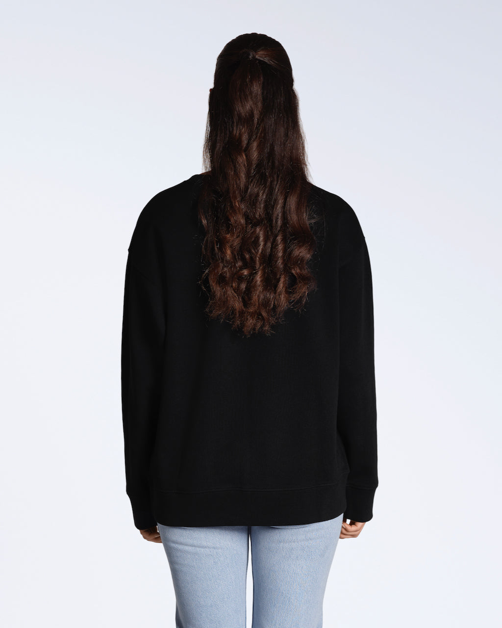 A woman facing away from the camera, modelling a plain black, oversized, organic cotton sweatshirt.