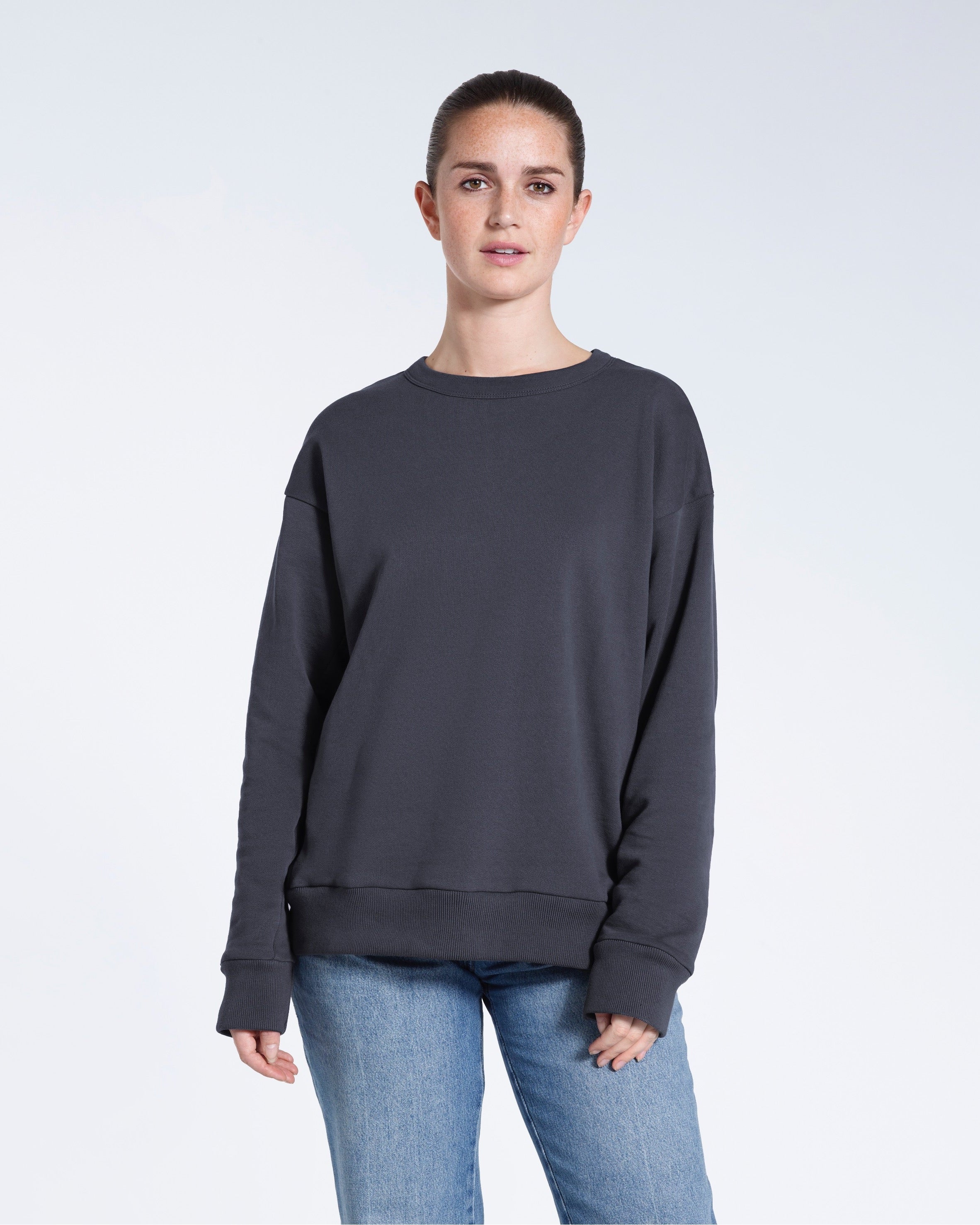 A woman looking into the camera with her arms by her side, modelling a plain dark grey oversized organic cotton sweatshirt.