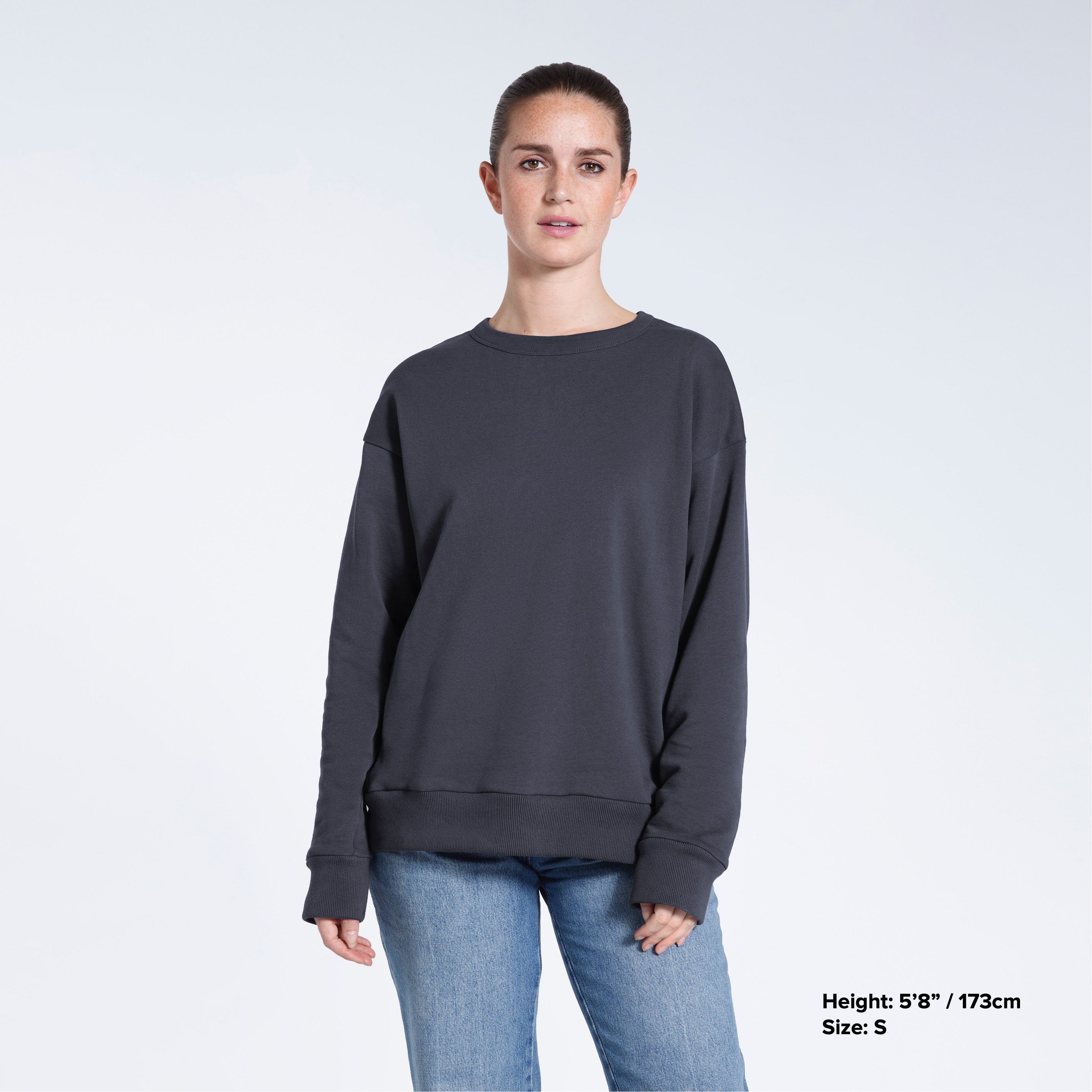 A woman looking into the camera with her arms by her side, modelling a plain dark grey oversized organic cotton sweatshirt.