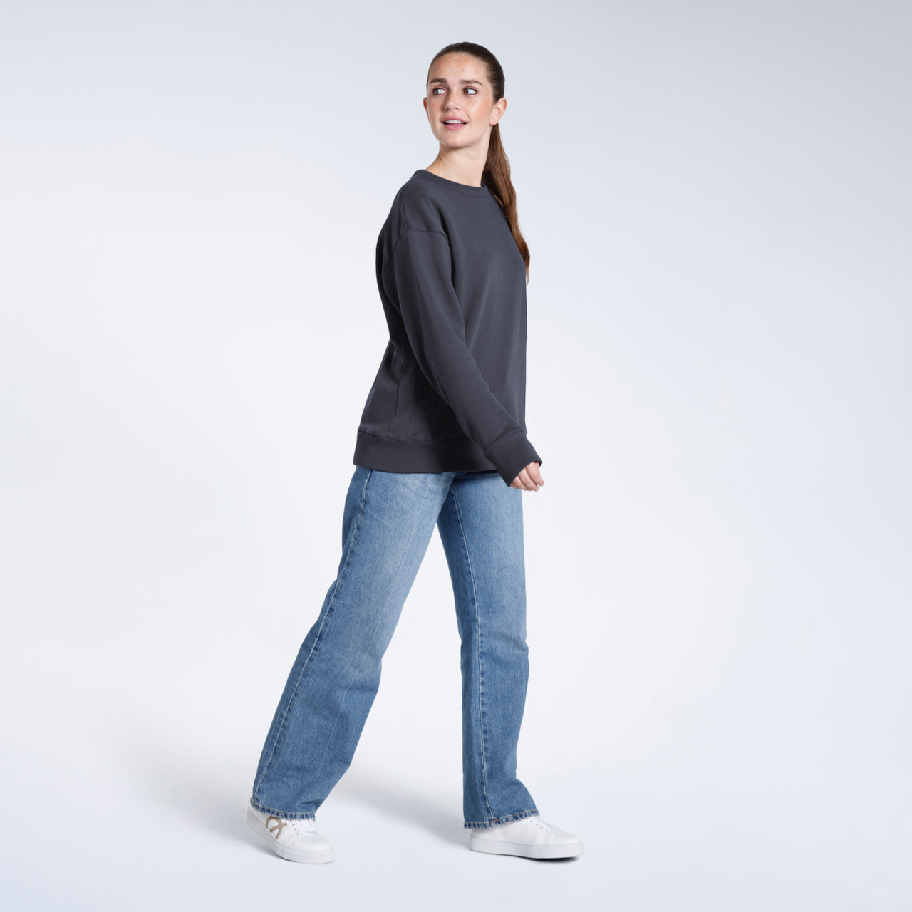 A full body photo of a woman looking over her shoulder, modelling a plain dark grey oversized organic cotton sweatshirt.
