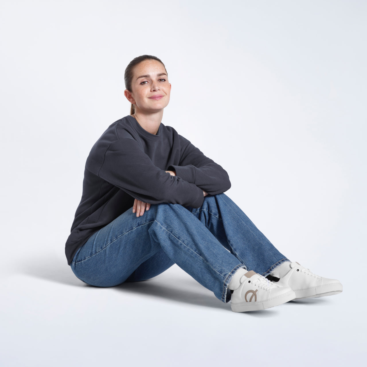 A woman sitting on the floor smiling, modelling a plain dark grey oversized organic cotton sweatshirt.