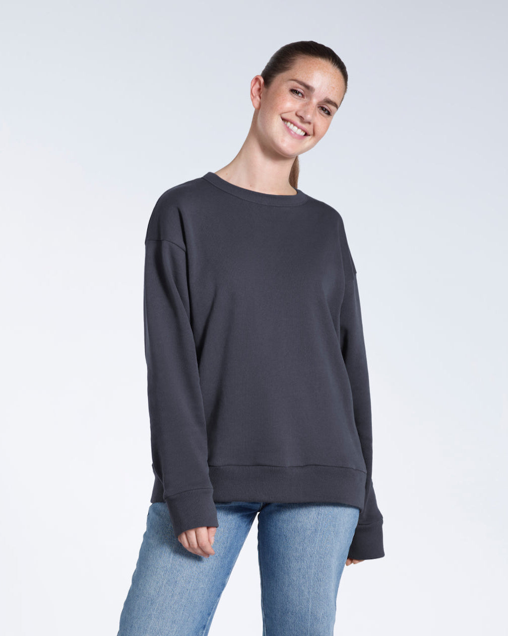 A woman smiling into the camera with her arms by her side, modelling a plain dark grey oversized organic cotton sweatshirt.