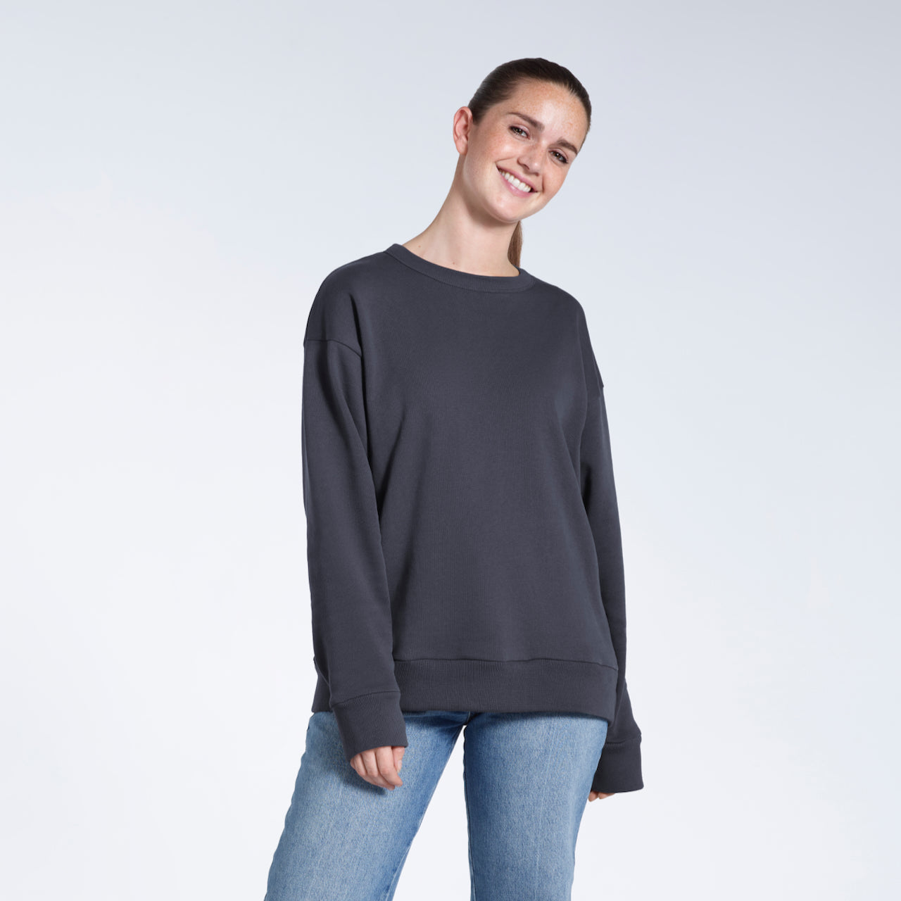 A woman smiling into the camera with her arms by her side, modelling a plain dark grey oversized organic cotton sweatshirt.