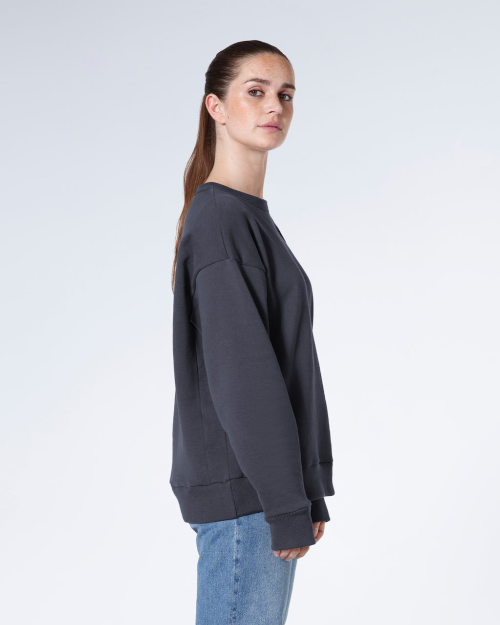 A woman facing side on looking into the camera, modelling a plain dark grey oversized organic cotton sweatshirt.