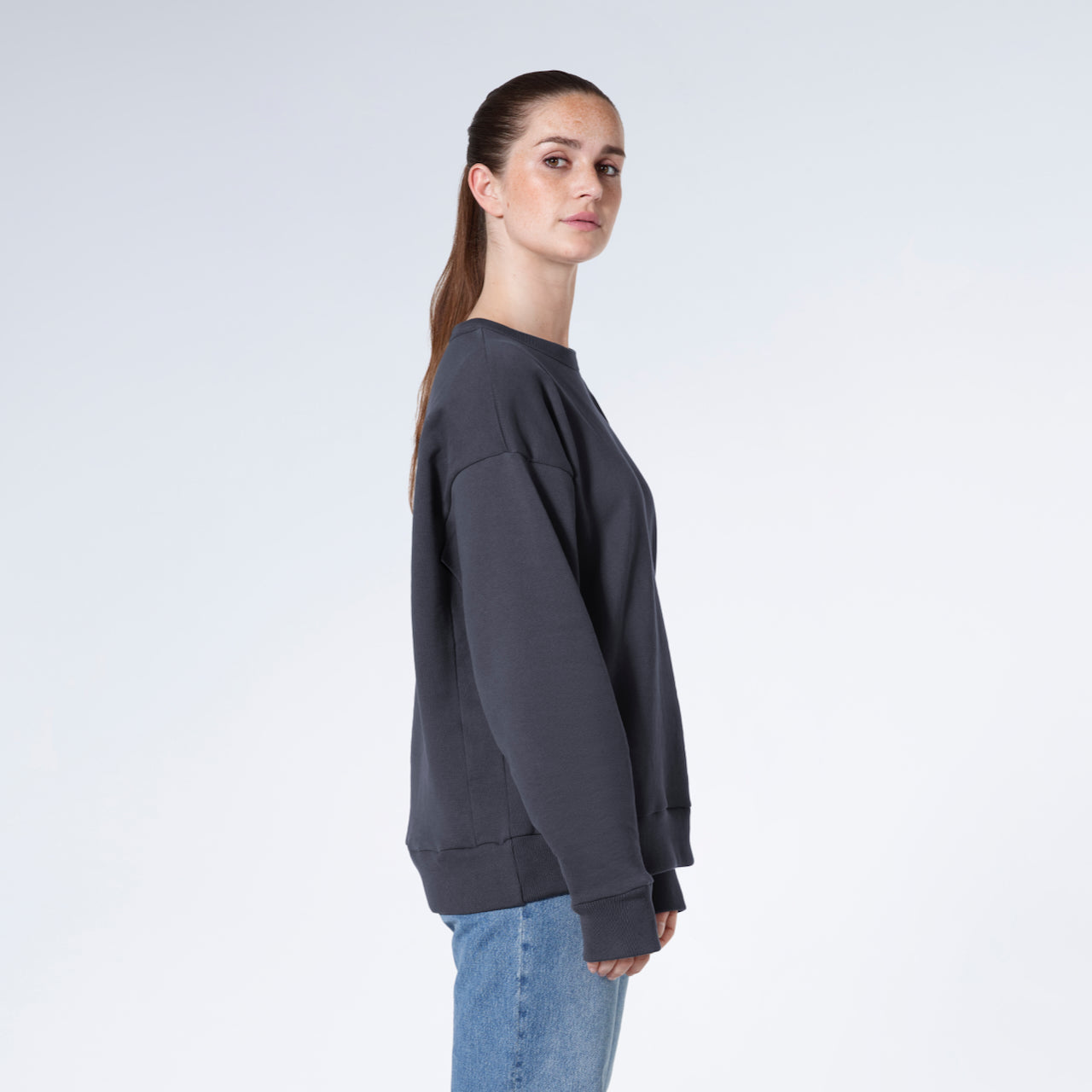 A woman facing side on looking into the camera, modelling a plain dark grey oversized organic cotton sweatshirt.