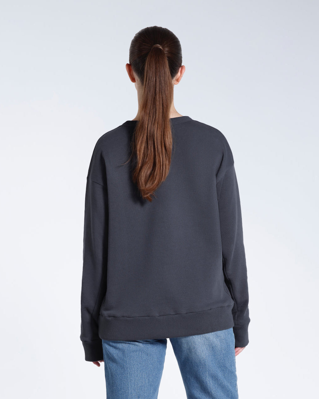 A woman facing away from the camera with her arms by her sides, modelling a plain dark grey oversized organic cotton sweatshirt.