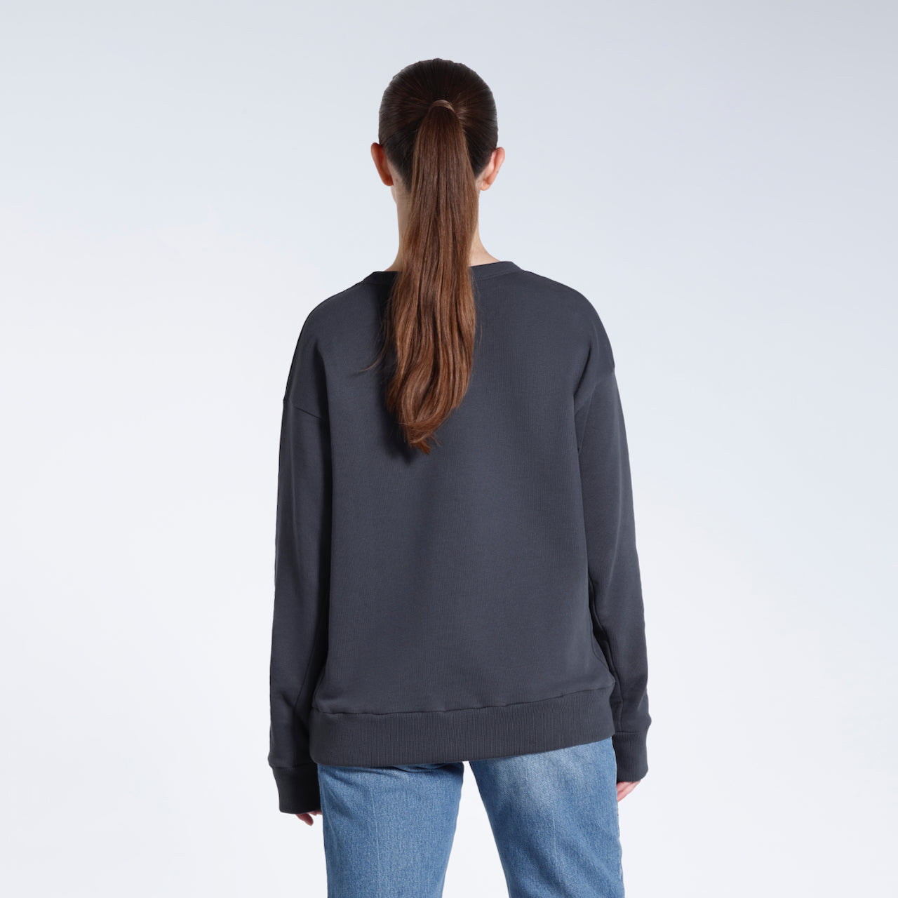 A woman facing away from the camera with her arms by her sides, modelling a plain dark grey oversized organic cotton sweatshirt.