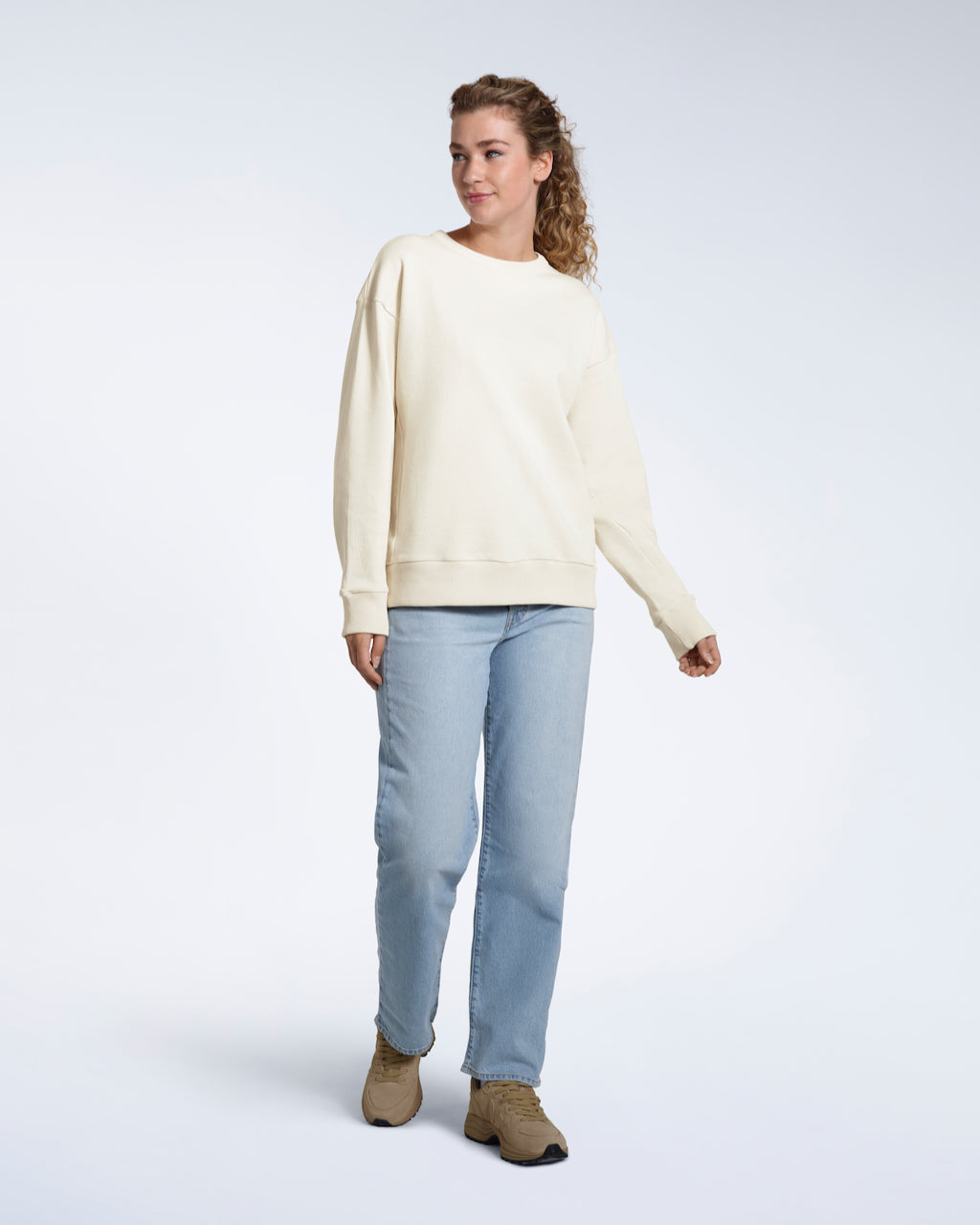 A full body photo of a woman smiling looking away from the camera, modelling a plain ecru oversized organic cotton sweatshirt.