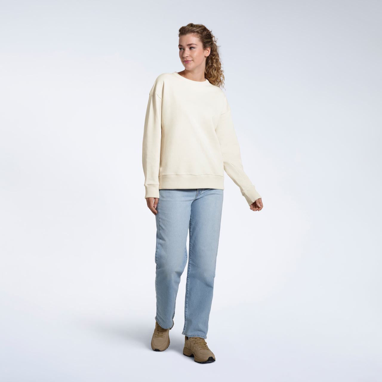 A full body photo of a woman smiling looking away from the camera, modelling a plain ecru oversized organic cotton sweatshirt.