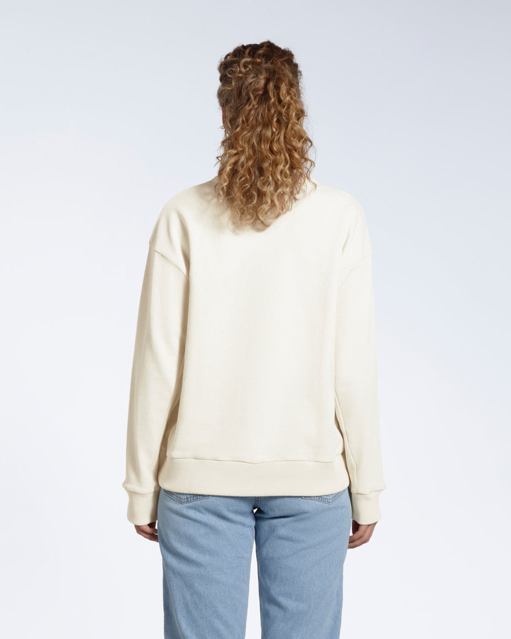 A woman facing away from the camera with her arms by her sides, modelling a plain ecru oversized organic cotton sweatshirt.