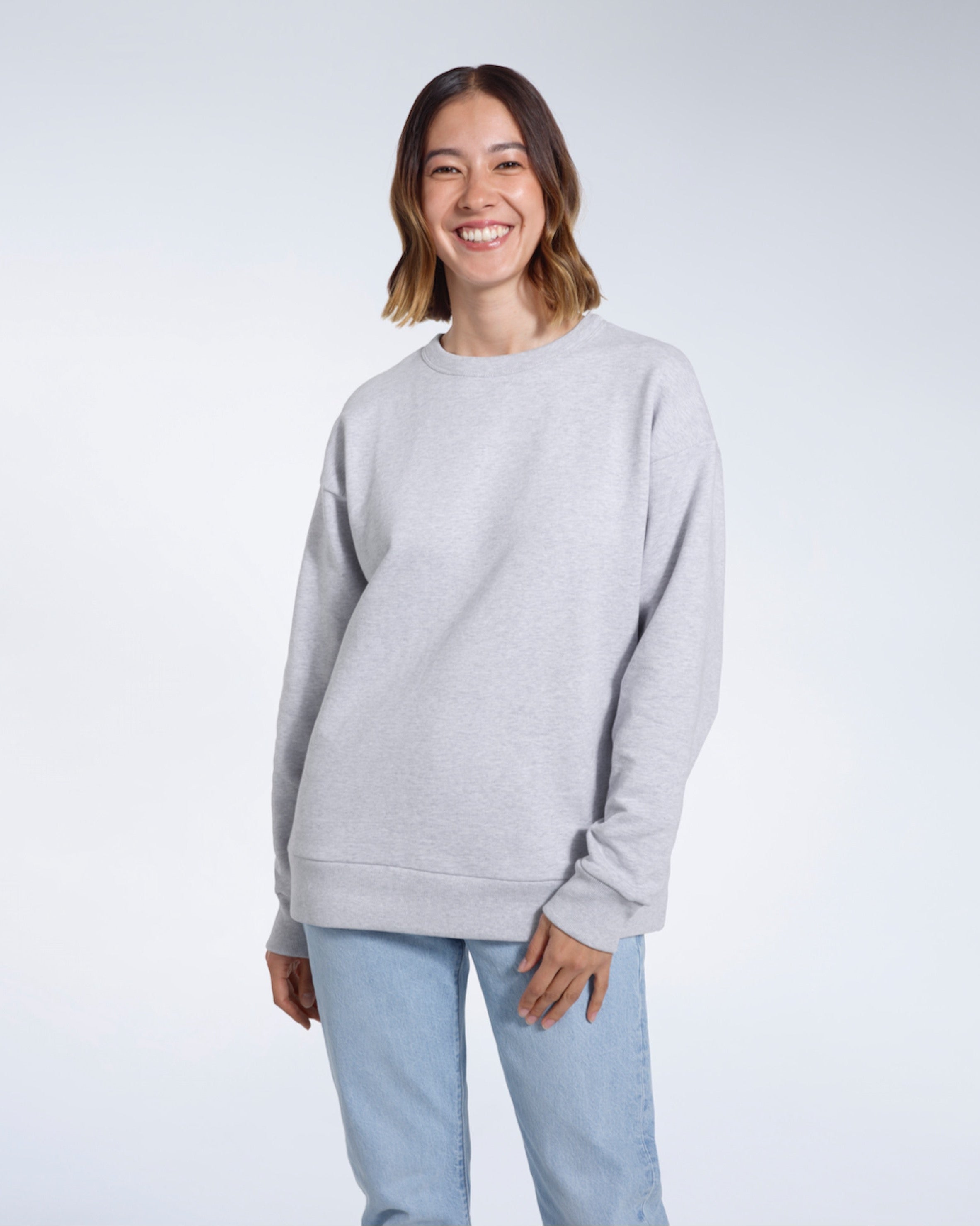 A woman smiling into the camera with her arms by her side, modelling a plain grey oversized organic cotton sweatshirt.