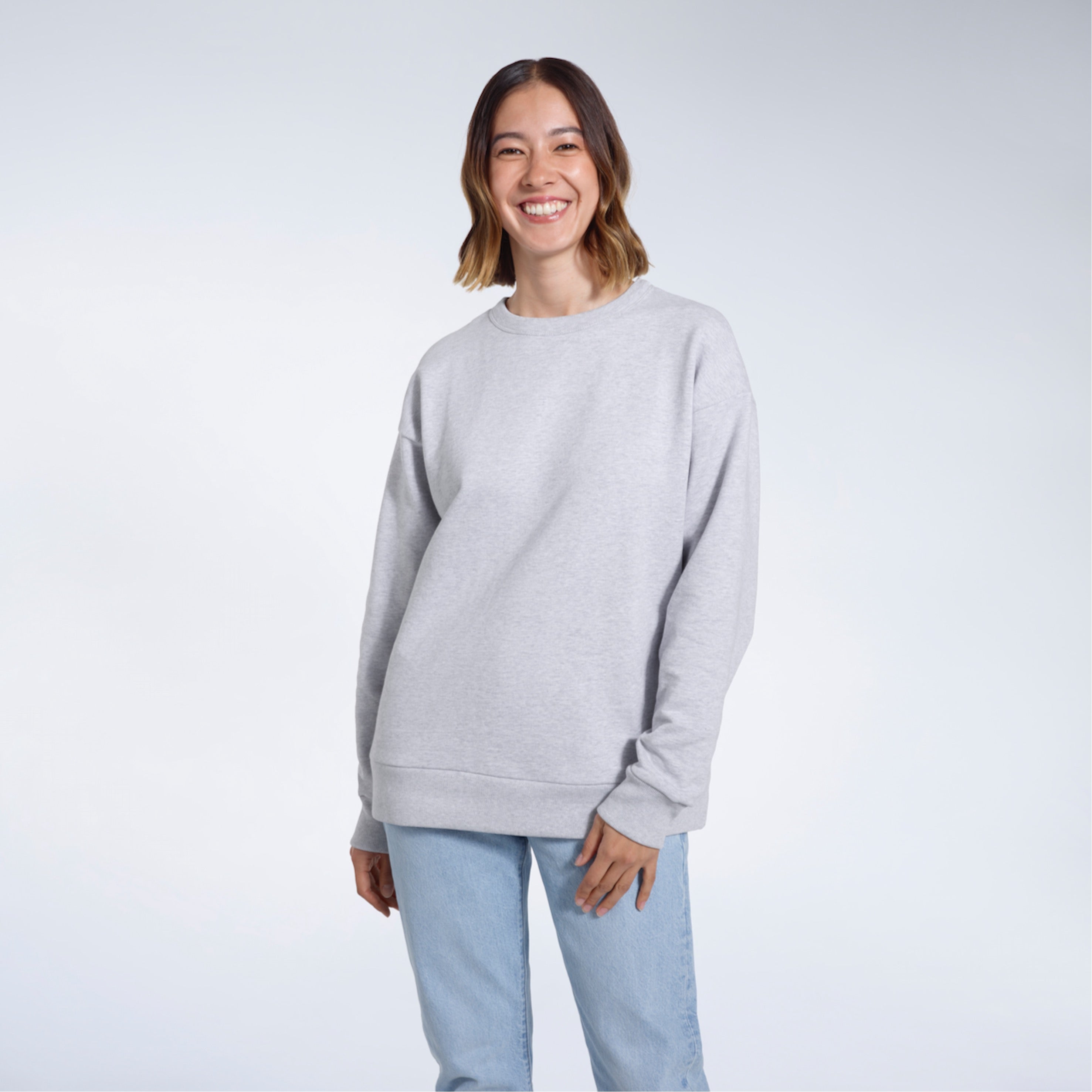 A woman smiling into the camera with her arms by her side, modelling a plain grey oversized organic cotton sweatshirt.