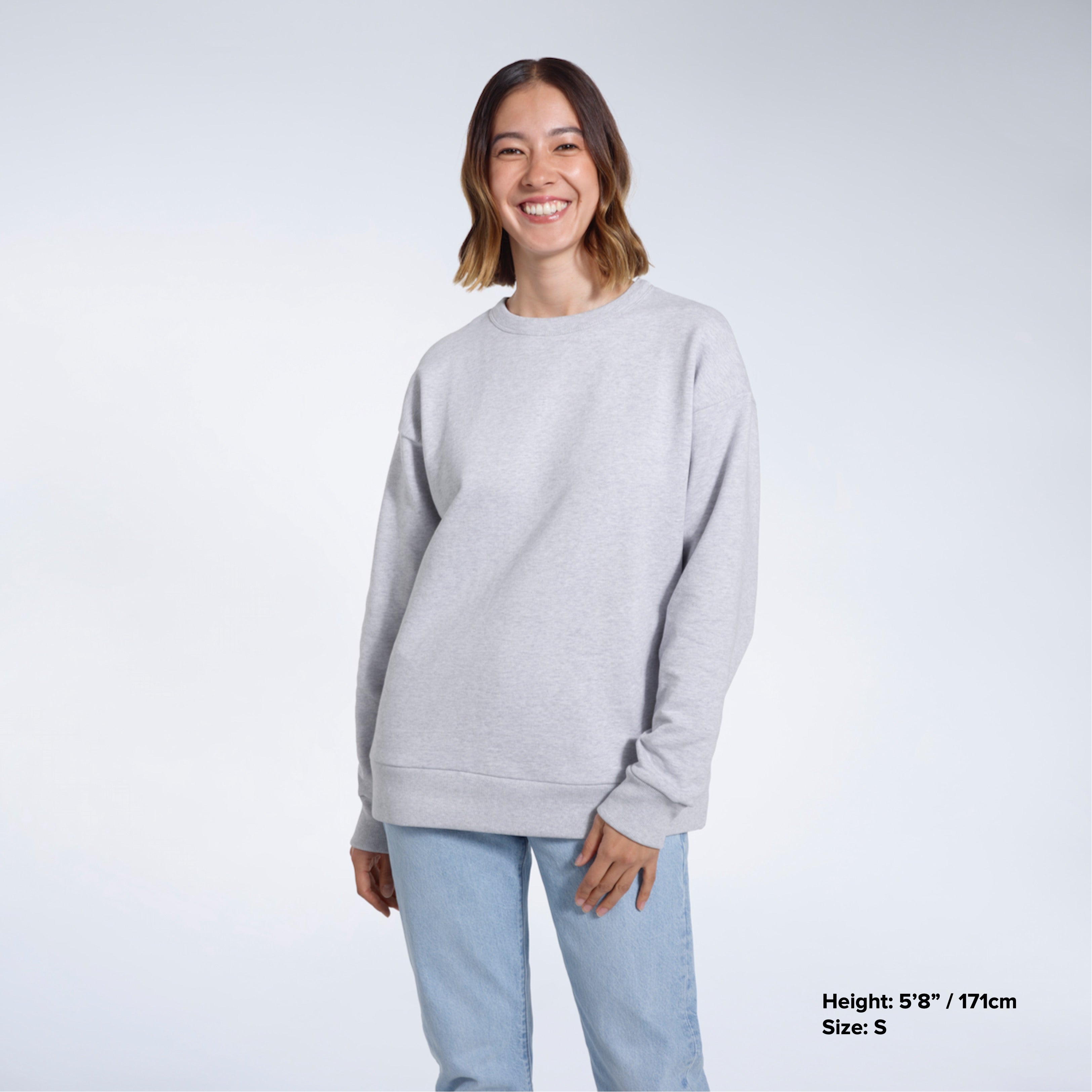 A woman smiling into the camera with her arms by her side, modelling a plain grey oversized organic cotton sweatshirt.