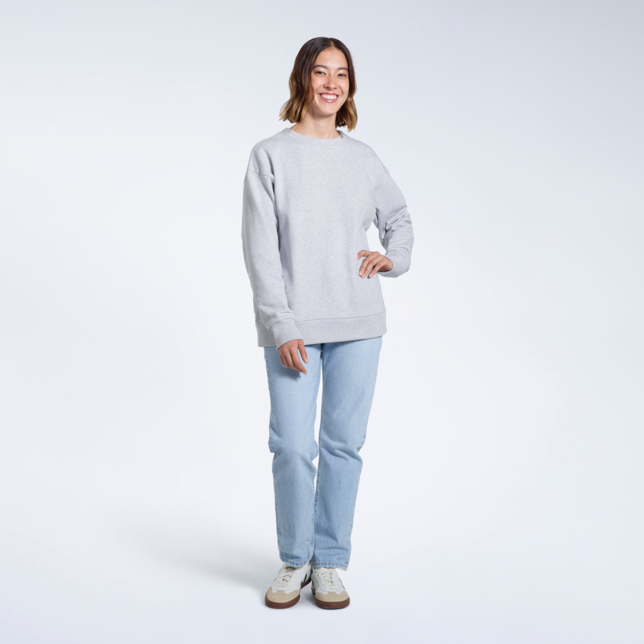 A full body photo of a woman smiling with her hand on her hip, modelling a plain grey oversized organic cotton sweatshirt.