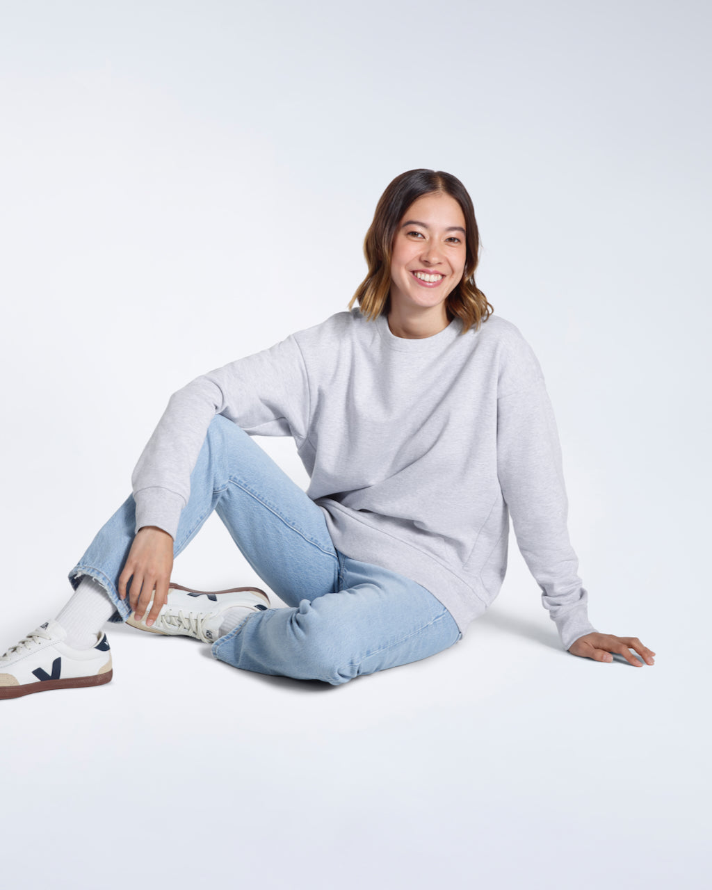 A woman sitting on the floor smiling, modelling a plain grey oversized organic cotton sweatshirt.