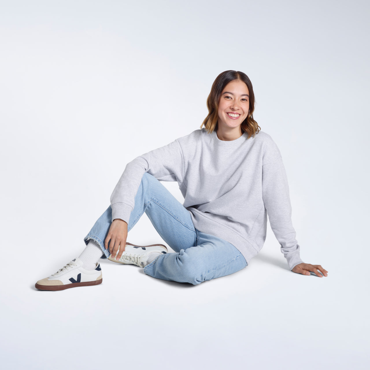 A woman sitting on the floor smiling, modelling a plain grey oversized organic cotton sweatshirt.