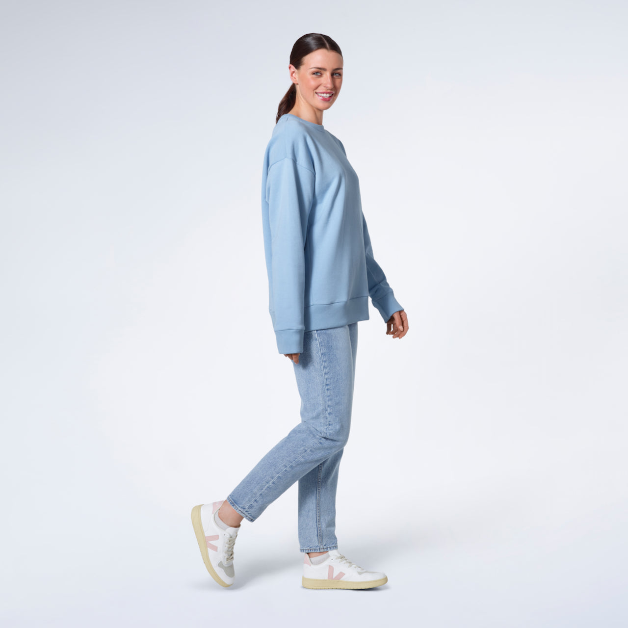 A full body photo of a woman walking and smiling into the camera, modelling a plain blue oversized organic cotton sweatshirt.