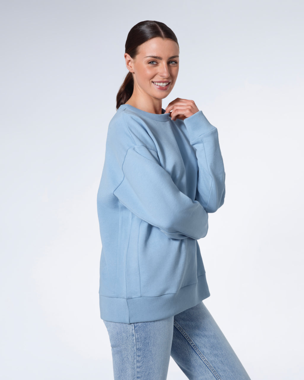 A woman smiling into the camera touching her collar, modelling a plain blue oversized organic cotton sweatshirt.