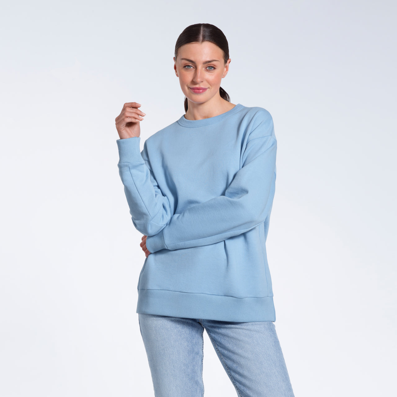 A woman smiling into the camera with her arm across her body, modelling a plain blue oversized organic cotton sweatshirt.