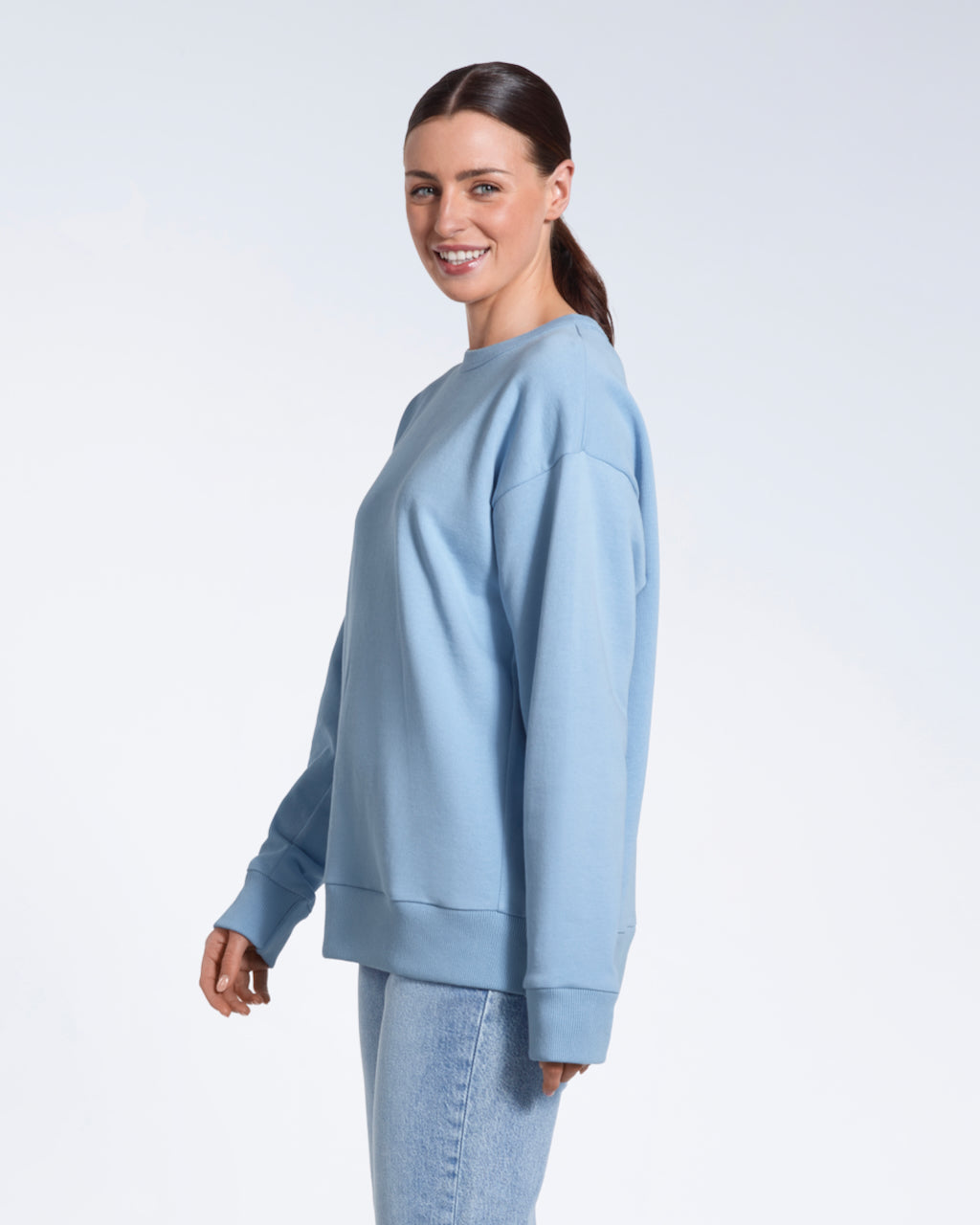 A woman walking and smiling into the camera, modelling a plain blue oversized organic cotton sweatshirt.