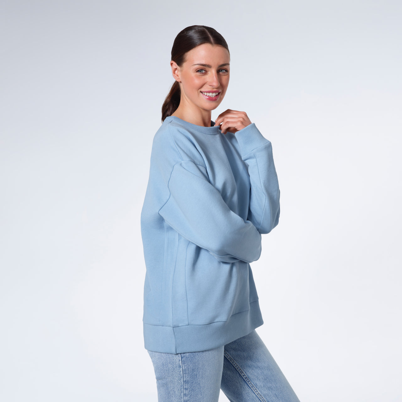 A woman smiling into the camera touching her collar, modelling a plain blue oversized organic cotton sweatshirt.