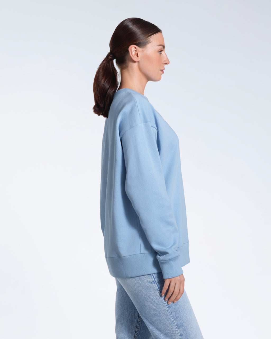 A woman facing side on with her arms by her side, modelling a plain blue oversized organic cotton sweatshirt.