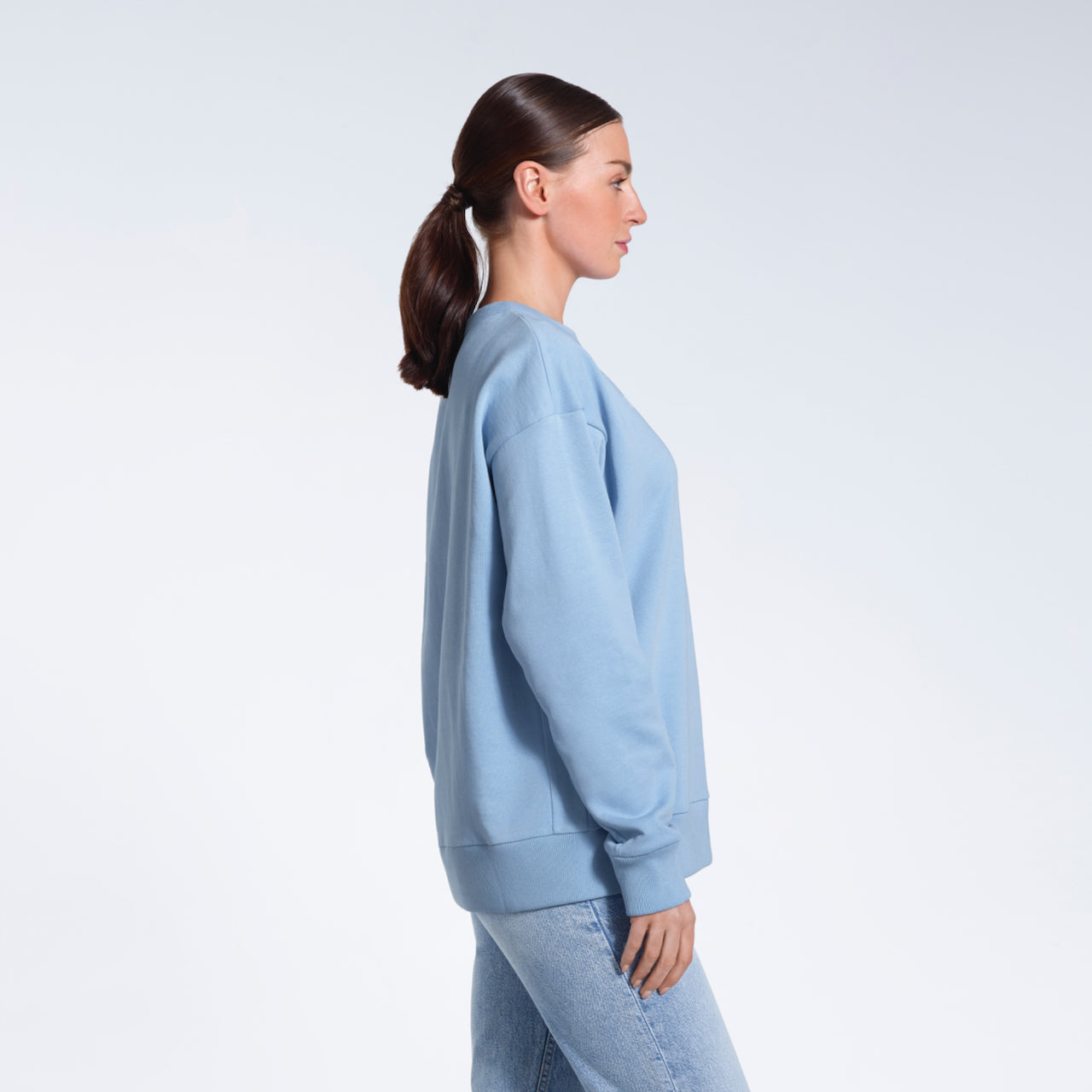 A woman facing side on with her arms by her side, modelling a plain blue oversized organic cotton sweatshirt.