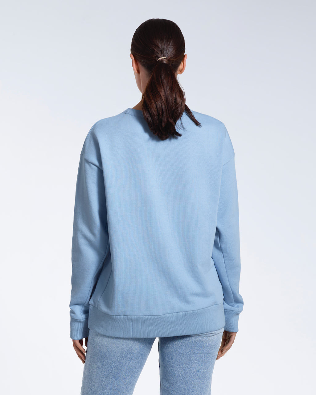 A woman facing away from the camera with her arms by her sides, modelling a plain blue oversized organic cotton sweatshirt.