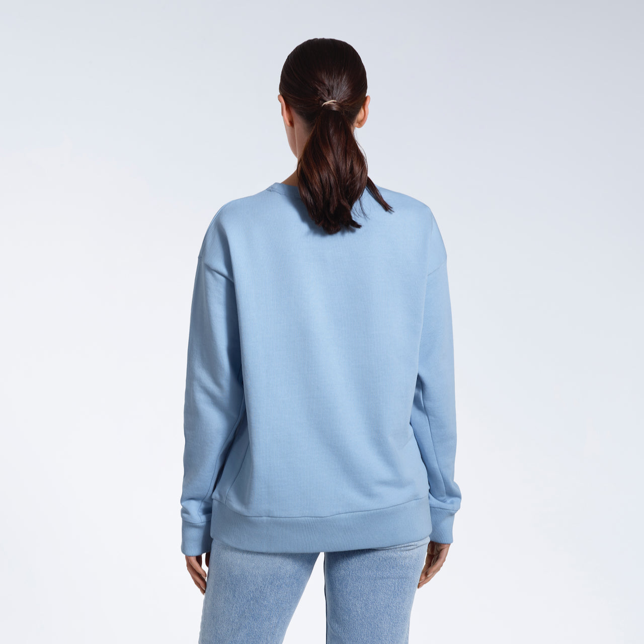 A woman facing away from the camera with her arms by her sides, modelling a plain blue oversized organic cotton sweatshirt.