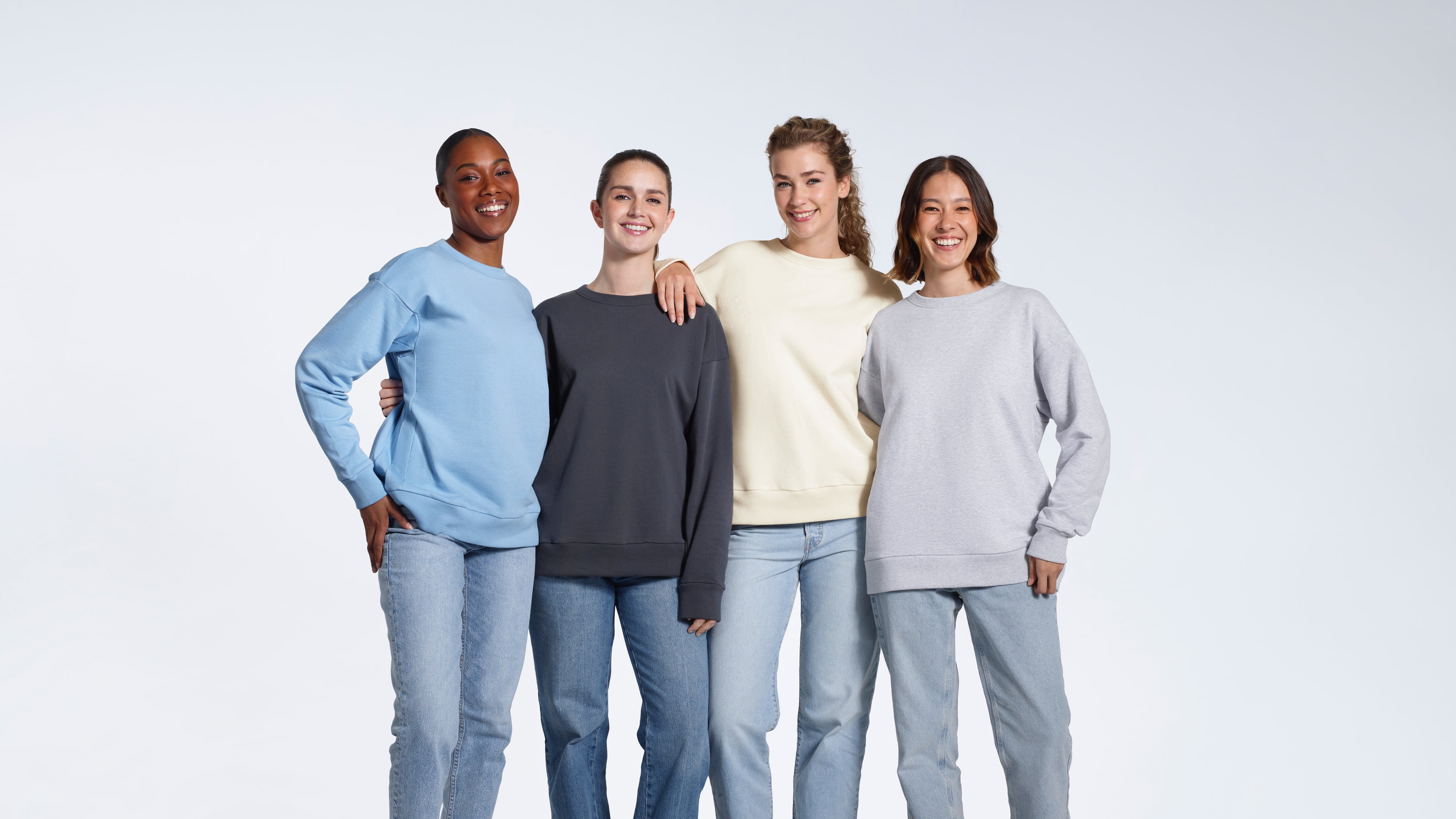 Four women smiling into the camera, modelling plain light blue, dark grey, ecru and grey coloured oversized organic cotton sweatshirts.