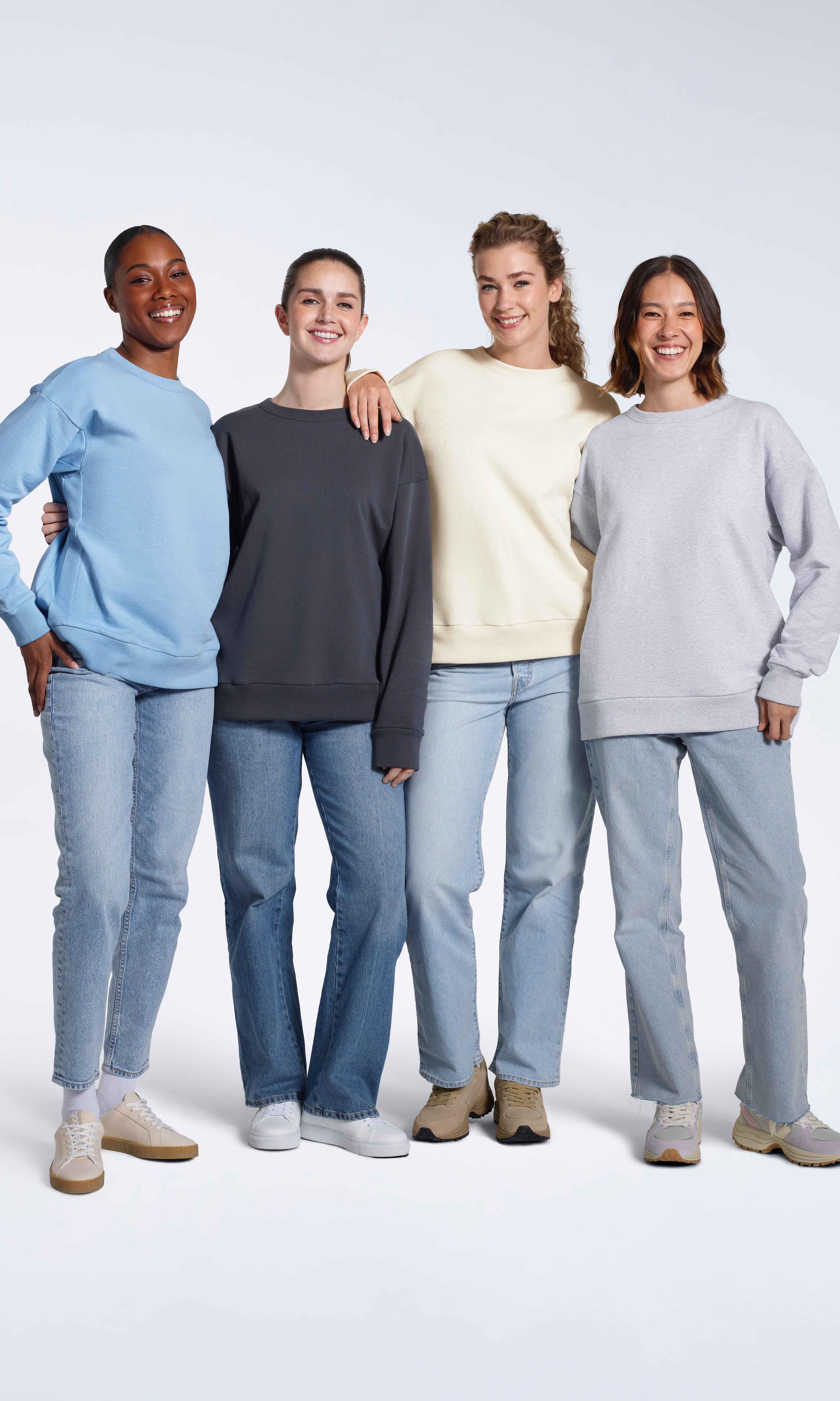 Four women smiling into the camera, modelling plain light blue, dark grey, ecru and grey coloured oversized organic cotton sweatshirts.