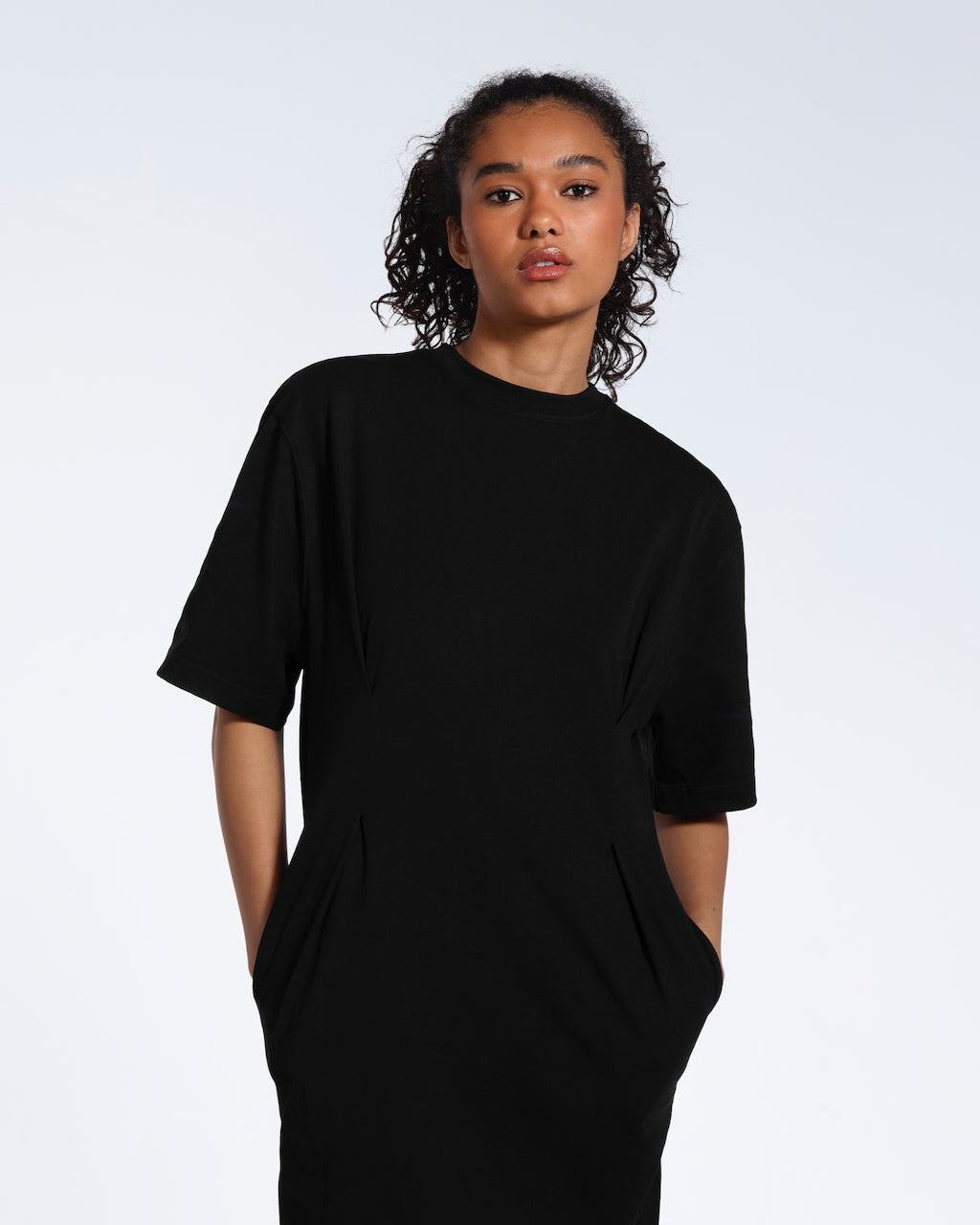 A women looking into the camera with her hands in her pockets, modelling a plain black, organic cotton midi t shirt dress.