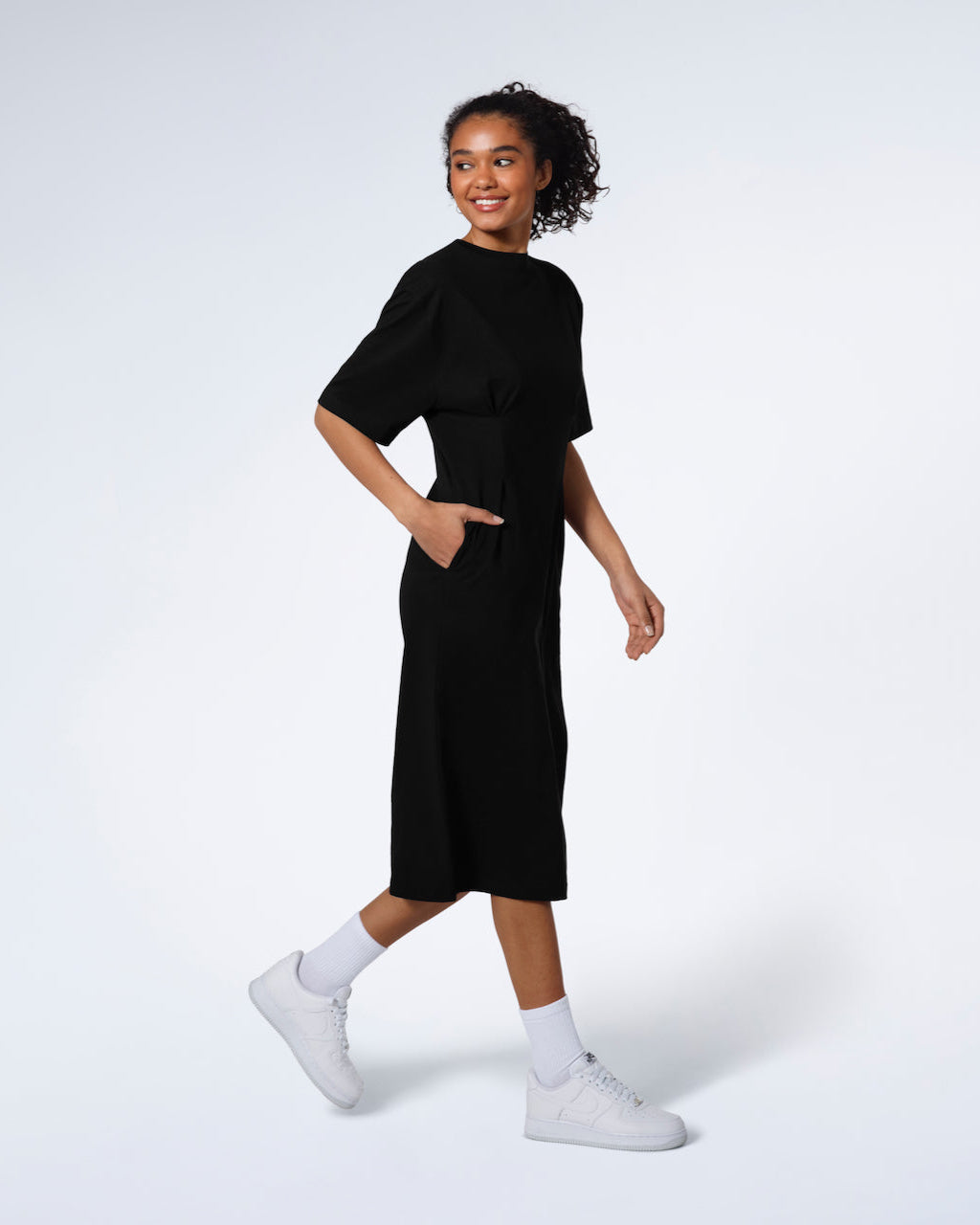 A women smiling and walking with her hand in her pocket, modelling a plain black, organic cotton midi t shirt dress.