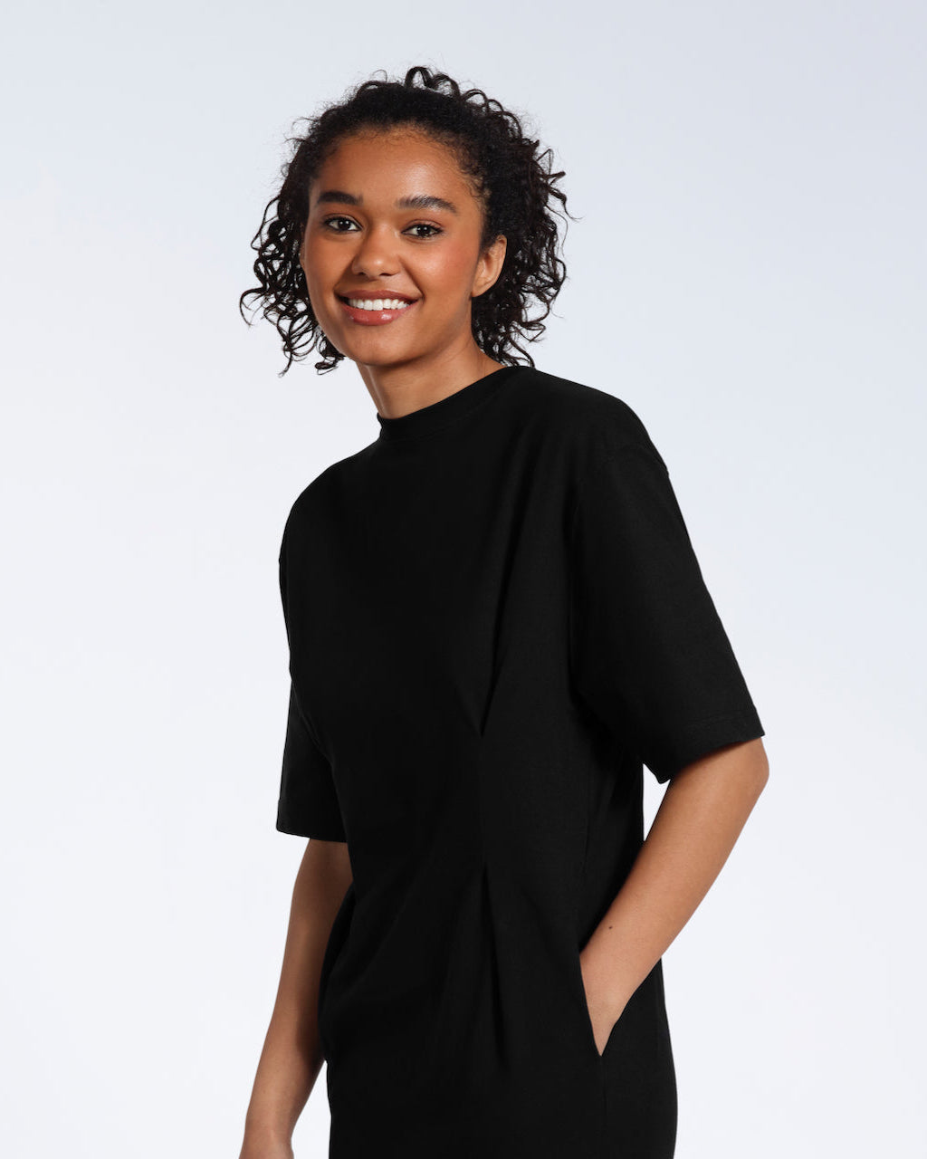 A women looking into the camera smiling with her hand in her pocket, modelling a plain black, organic cotton midi t shirt dress.